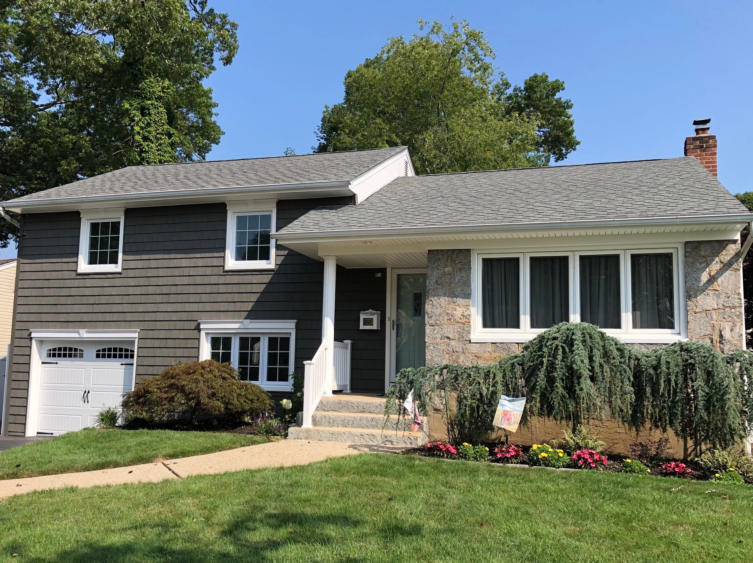 A two-story house with dark gray siding, white trim, and a stone facade on part of the front. The house has a gable roof with gray shingles, a white front porch with stairs, and a white garage door. There are windows on the front, and a brick chimney