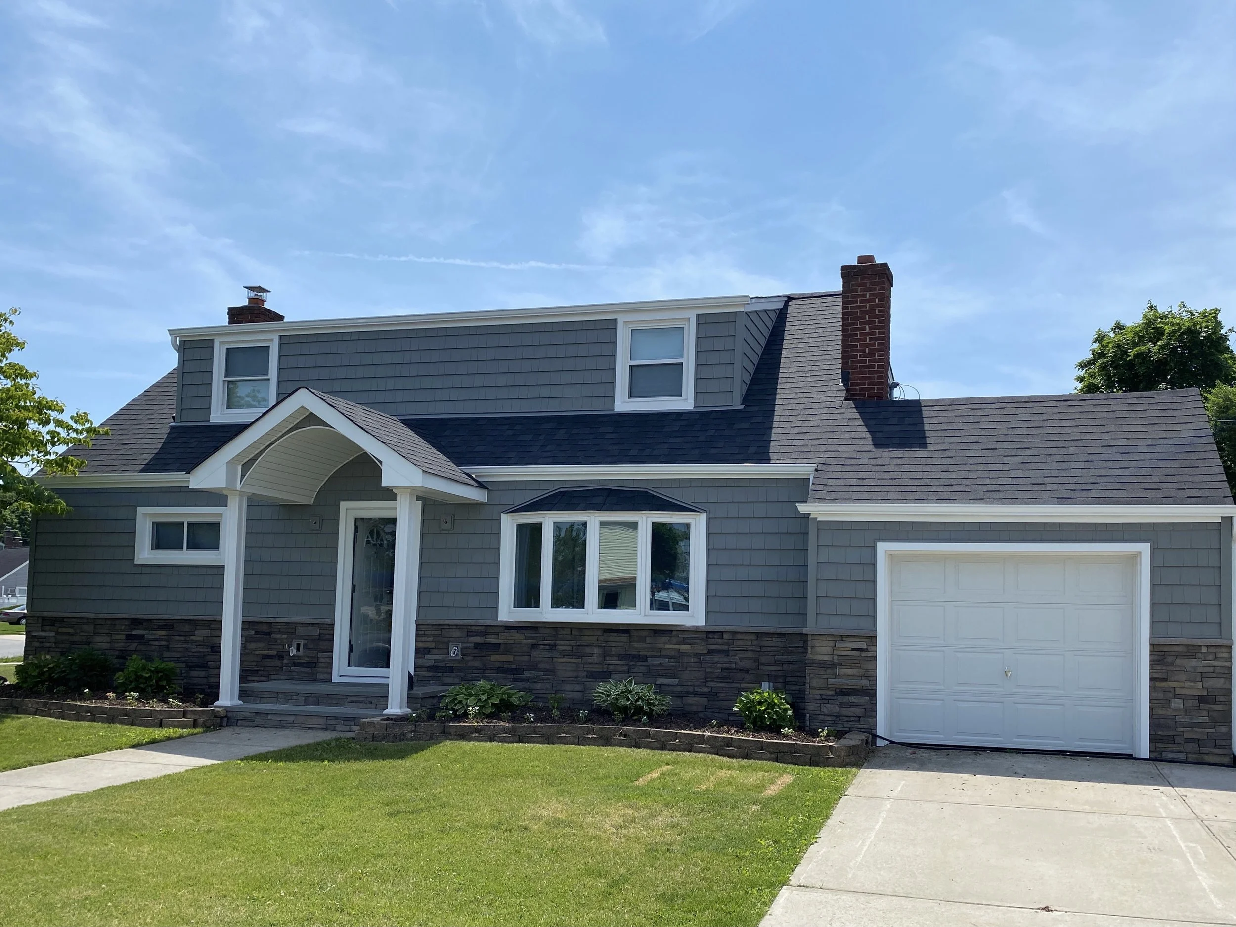 A two-story house with gray siding, a brick chimney, white trim, a front porch, and a white garage door, with a well-maintained lawn and a sidewalk in front, under a blue sky with some clouds.