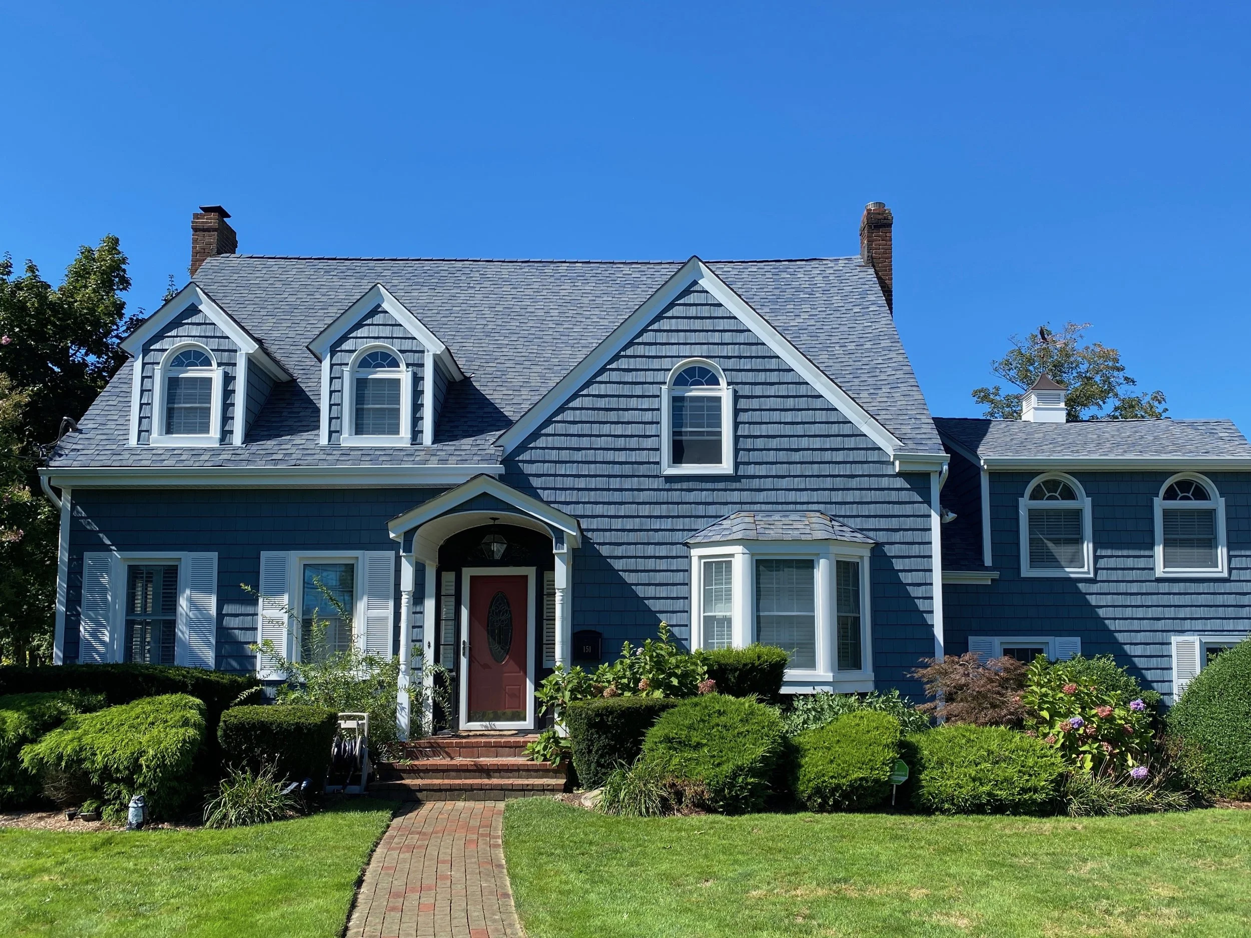 A blue house with multiple gables and arched windows, surrounded by a well-maintained garden and a brick pathway leading to the front door, under a clear blue sky.