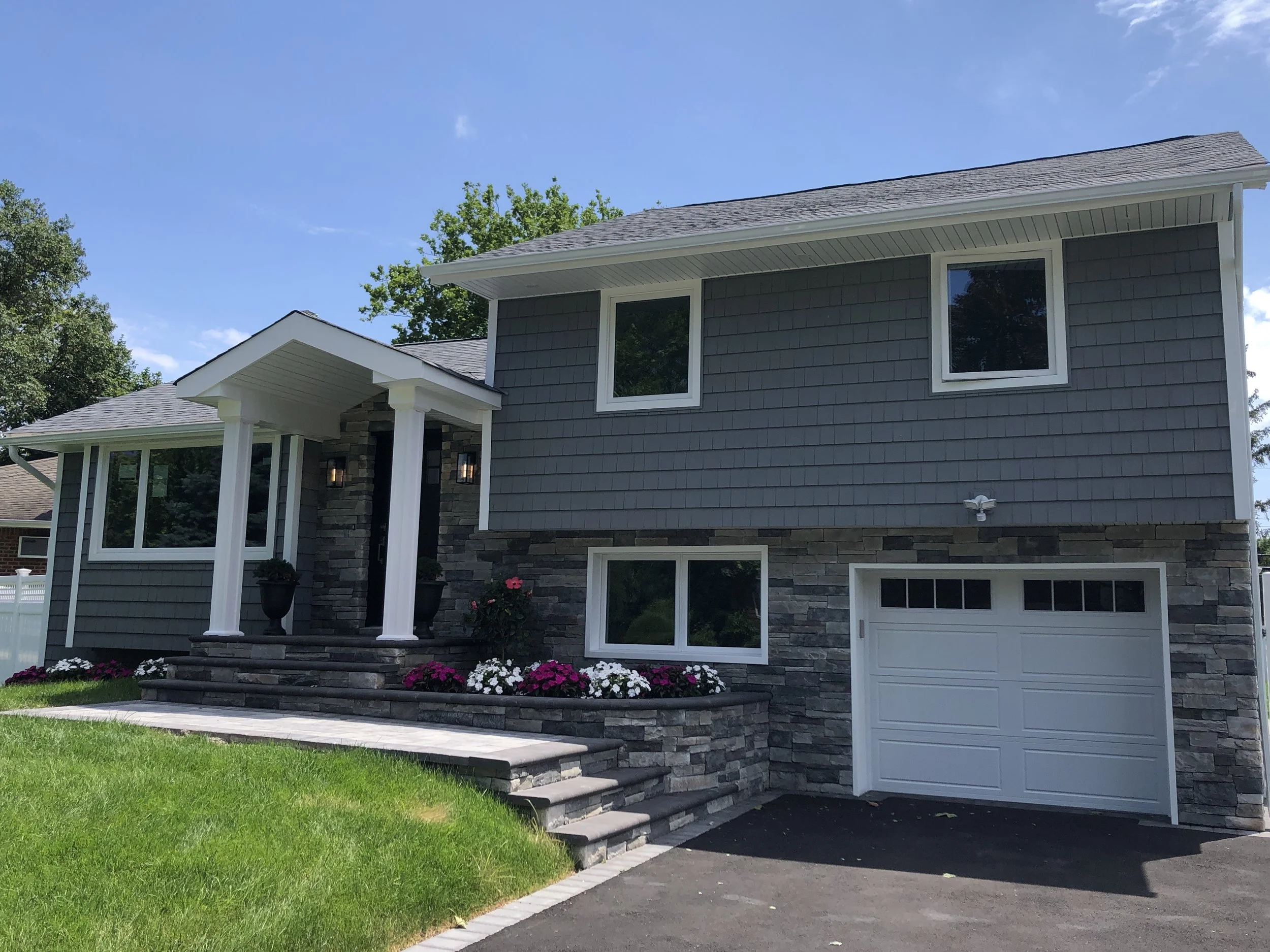 A modern two-story house with dark gray siding, stone accents, multiple windows, a white garage door, and a front porch with steps leading up to the entrance. Green lawn and colorful flowers in front, with trees and a blue sky in the background.