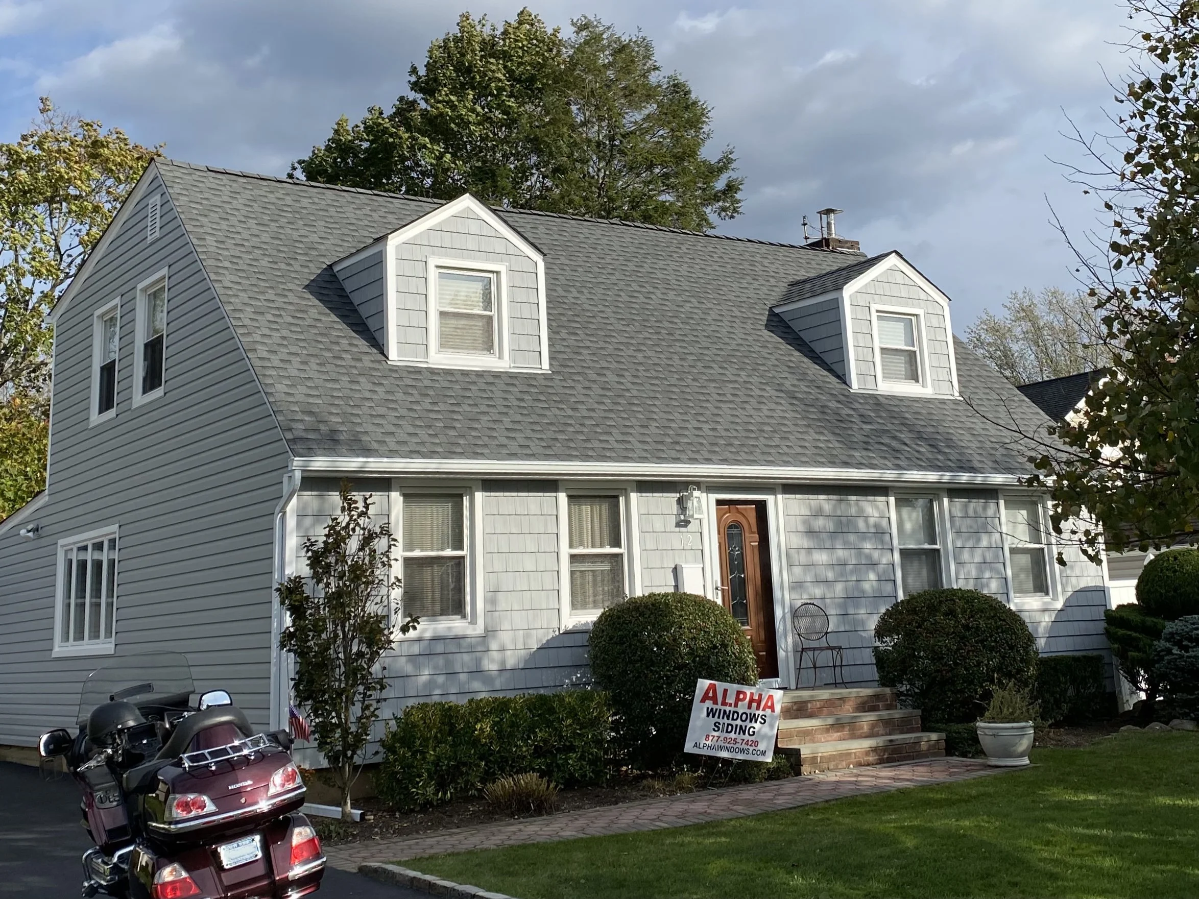 A house with light gray siding and two small dormer windows on the roof. There is a brick pathway leading to the front door, surrounded by bushes and a lawn. In the foreground, there are parked motorcycles. A sign in front advertises window siding se