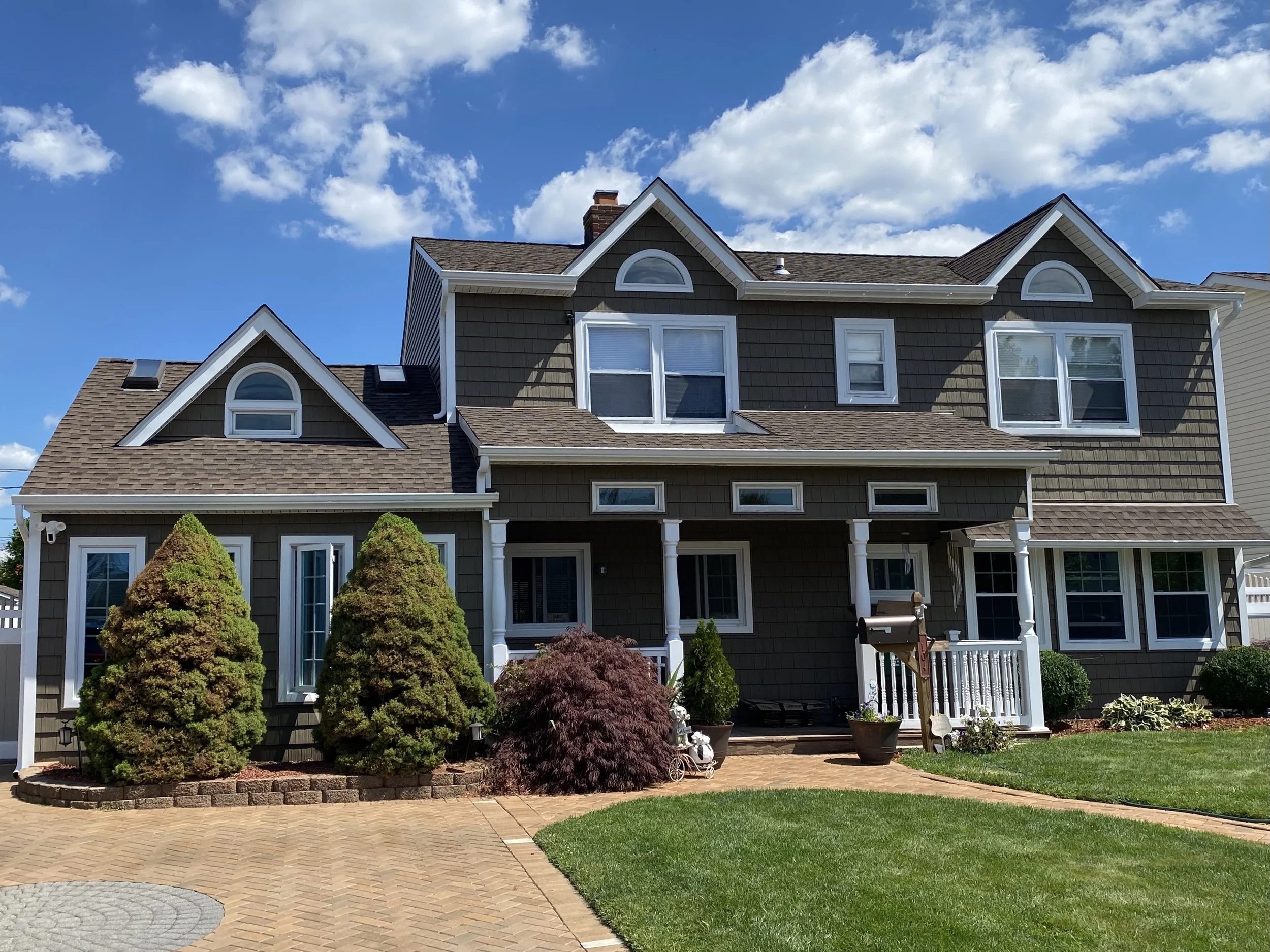 Front view of a large, multi-story house with gray siding, multiple gabled roofs, and several windows. The sky is blue with white clouds, and the house has a well-maintained yard with various shrubs and a brick patio.