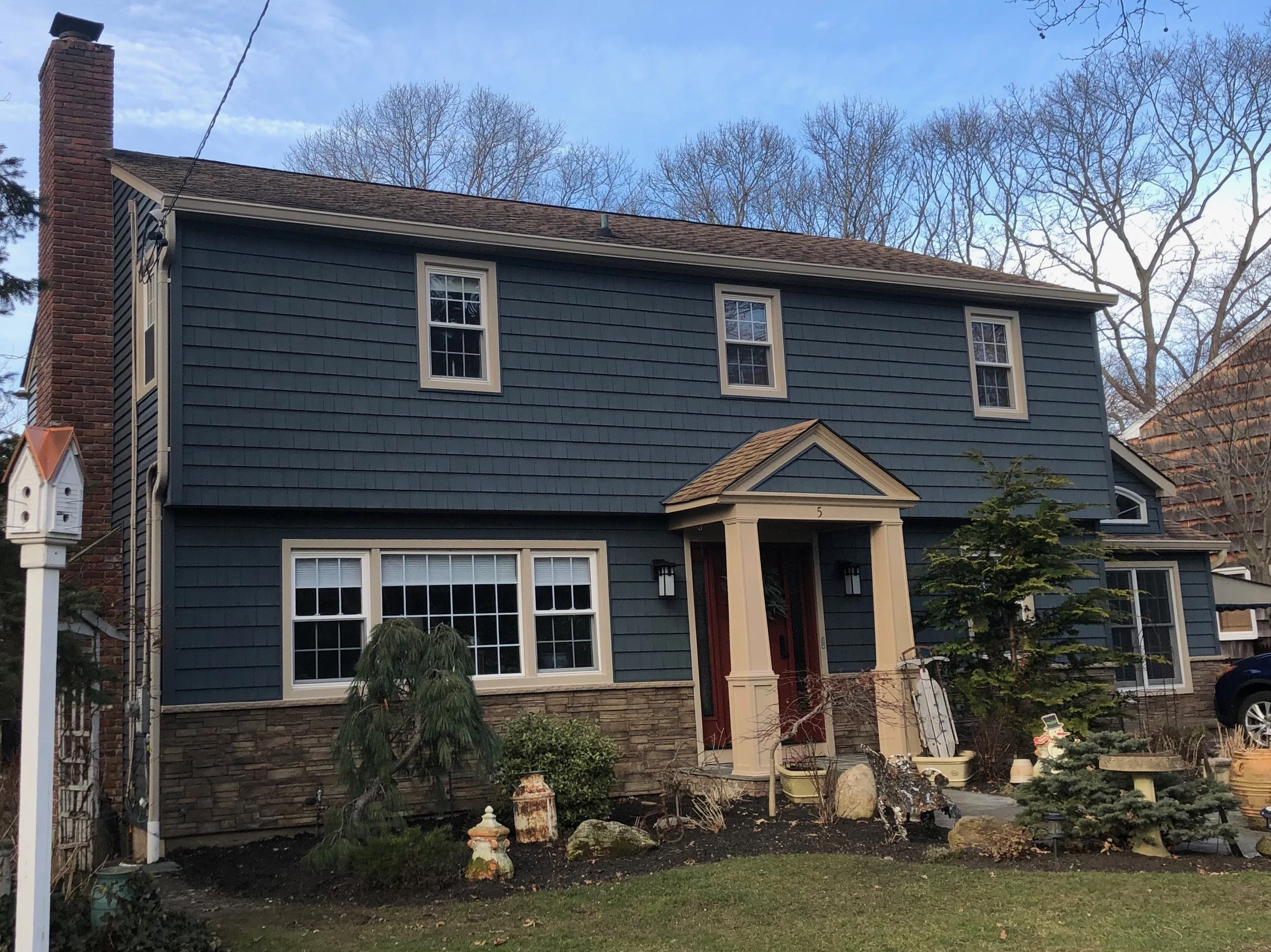 The photo shows the back of a two-story house with gray shingle siding, a brick chimney on the left, and multiple white-framed windows. The front yard features small trees, bushes, some garden statues, and a variety of plants. The sky is clear with a