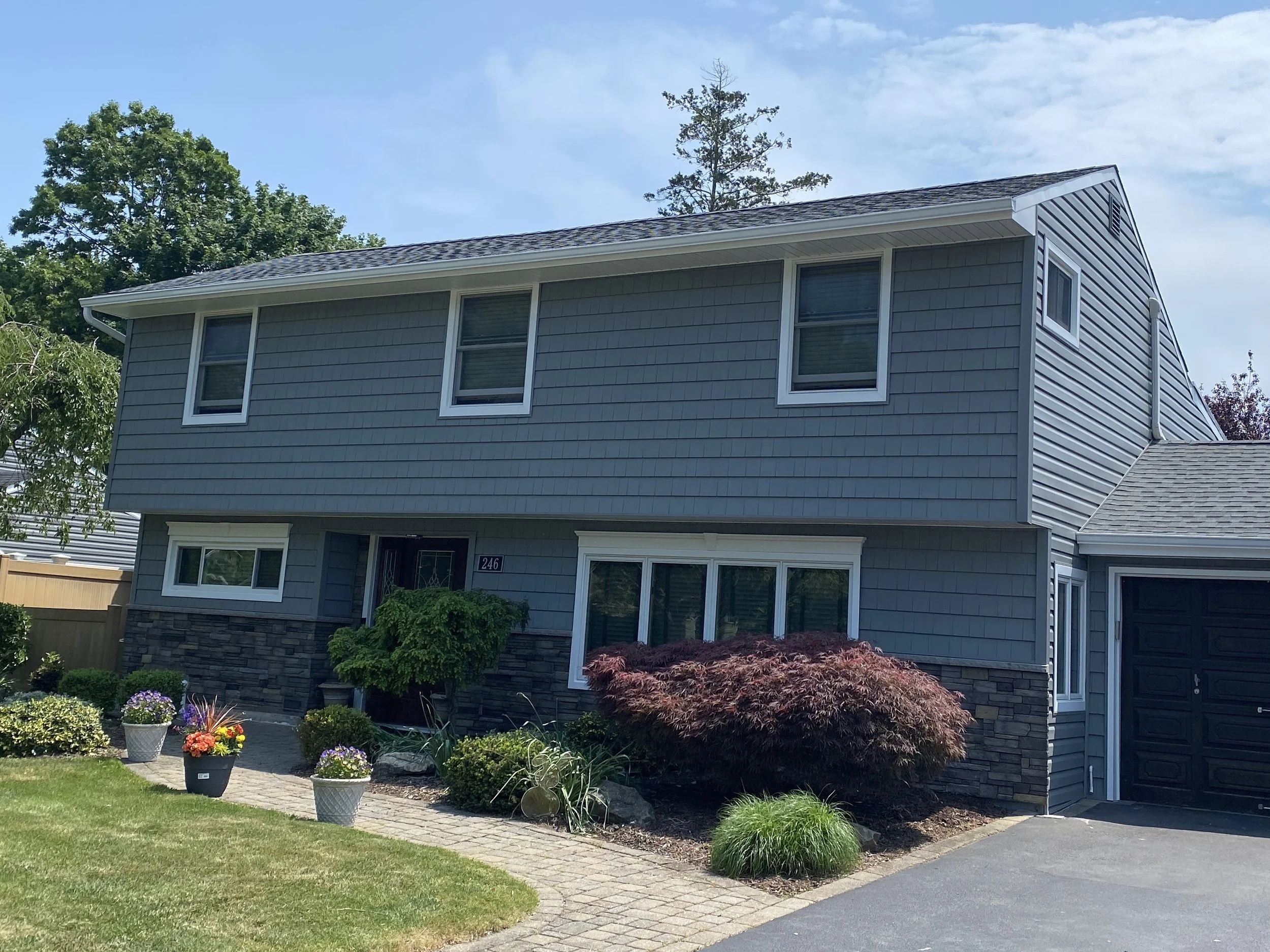 A two-story house with blue siding, gray shingle roof, and several double-pane windows. The front yard has a landscaped garden with potted flowers, shrubs, and small trees, and a paved walkway leading to the entrance.