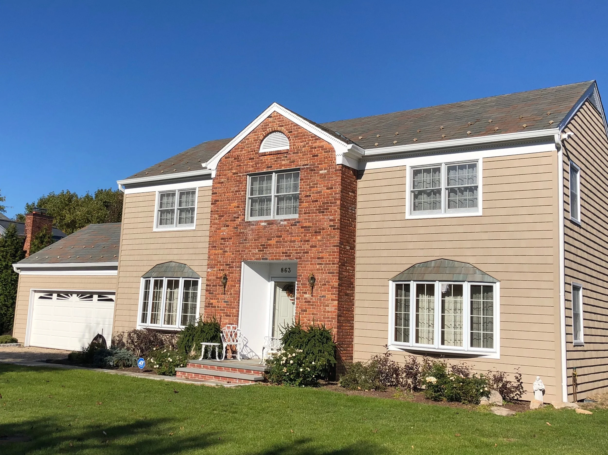 A two-story house with beige siding, a brick facade on the upper part, white-framed windows, a white front door, and a white garage door. The house has a sloped roof with shingles, a small front porch with brick steps, a lawn with some bushes and flo