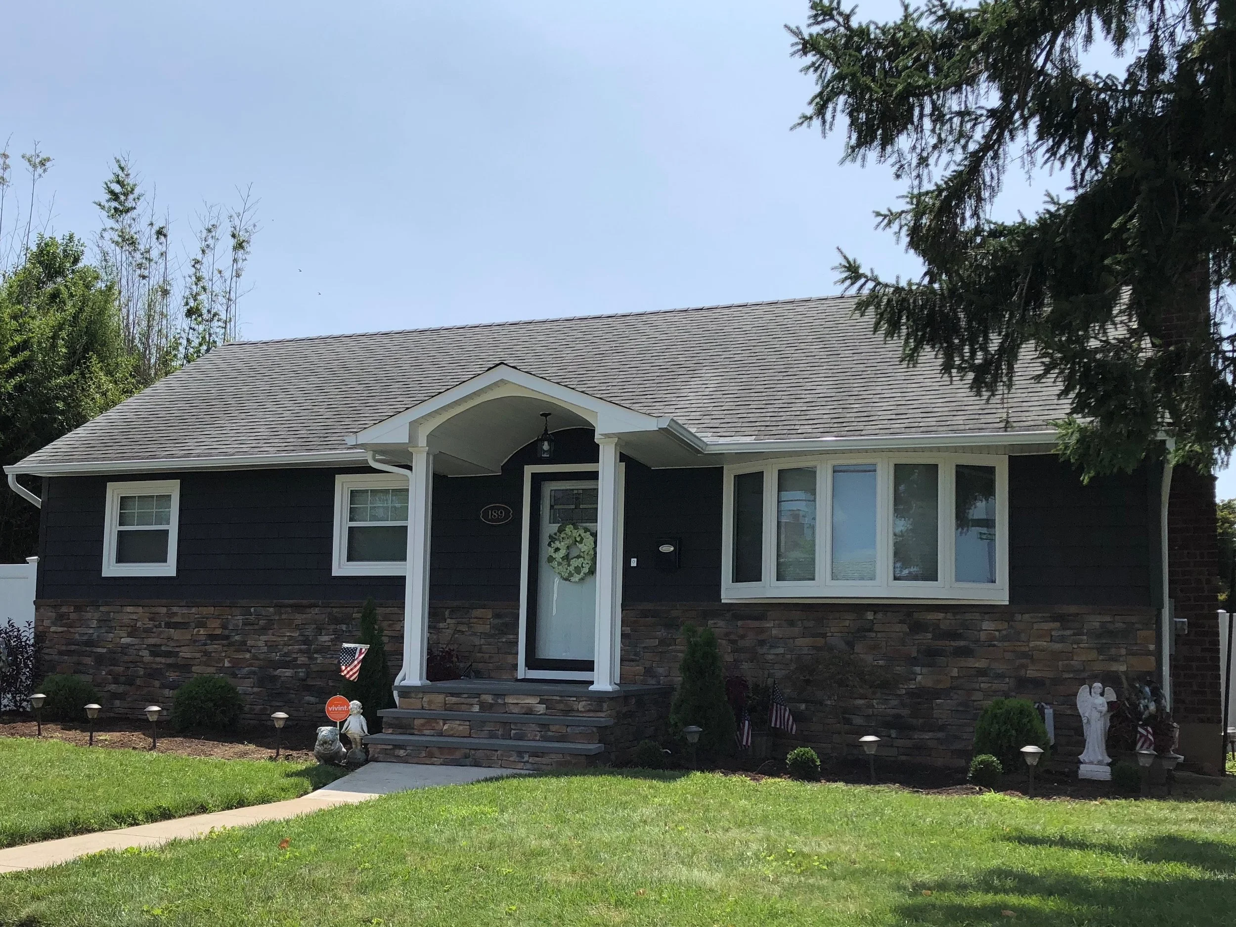 A house with a dark exterior, stone foundation, and a front porch decorated with a wreath and American flags. The house has a sloped roof and large windows, with a neatly maintained front yard and sidewalk leading to the entrance.