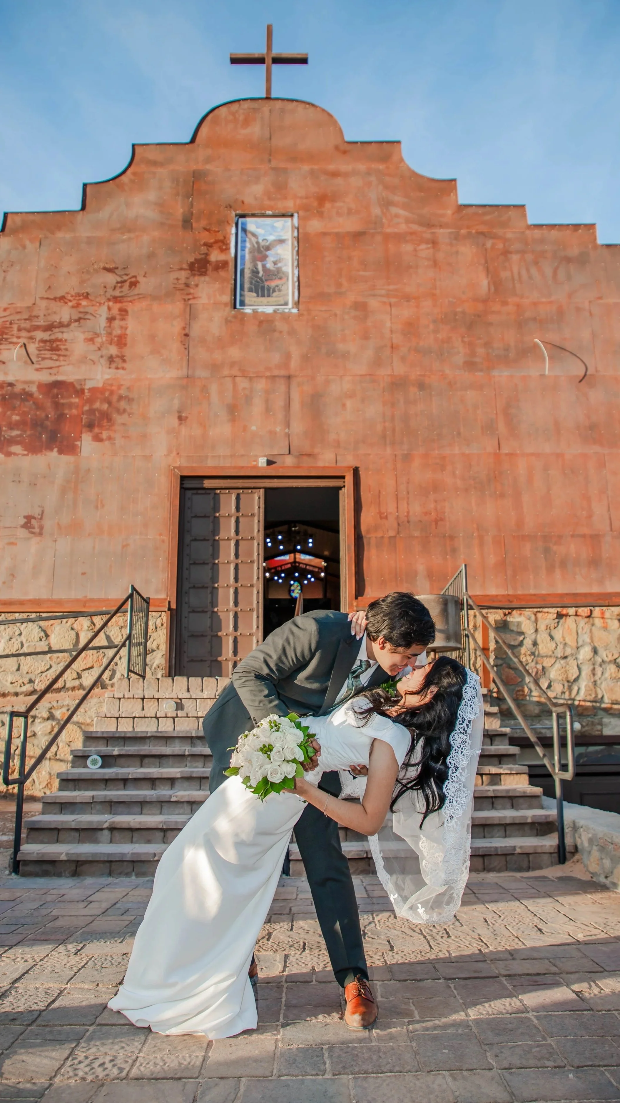 A bride and groom sharing a romantic moment on the steps of an El Paso church, with the groom leaning over the bride, who is holding a bouquet of white flowers, during their wedding celebration.