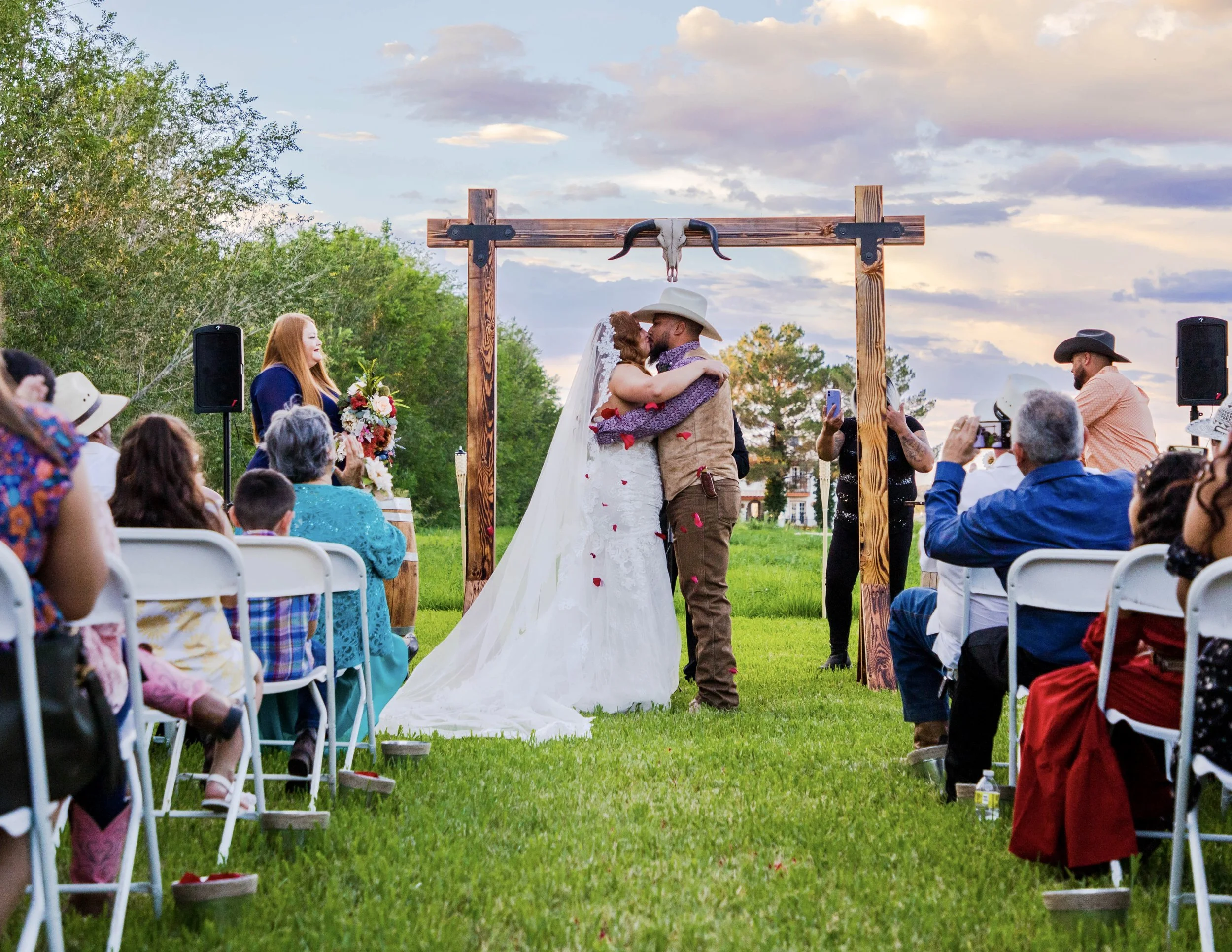 A bride and groom kissing under a wooden arch at an outdoor wedding ceremony during sunset, with guests seated on white chairs and trees in the background.