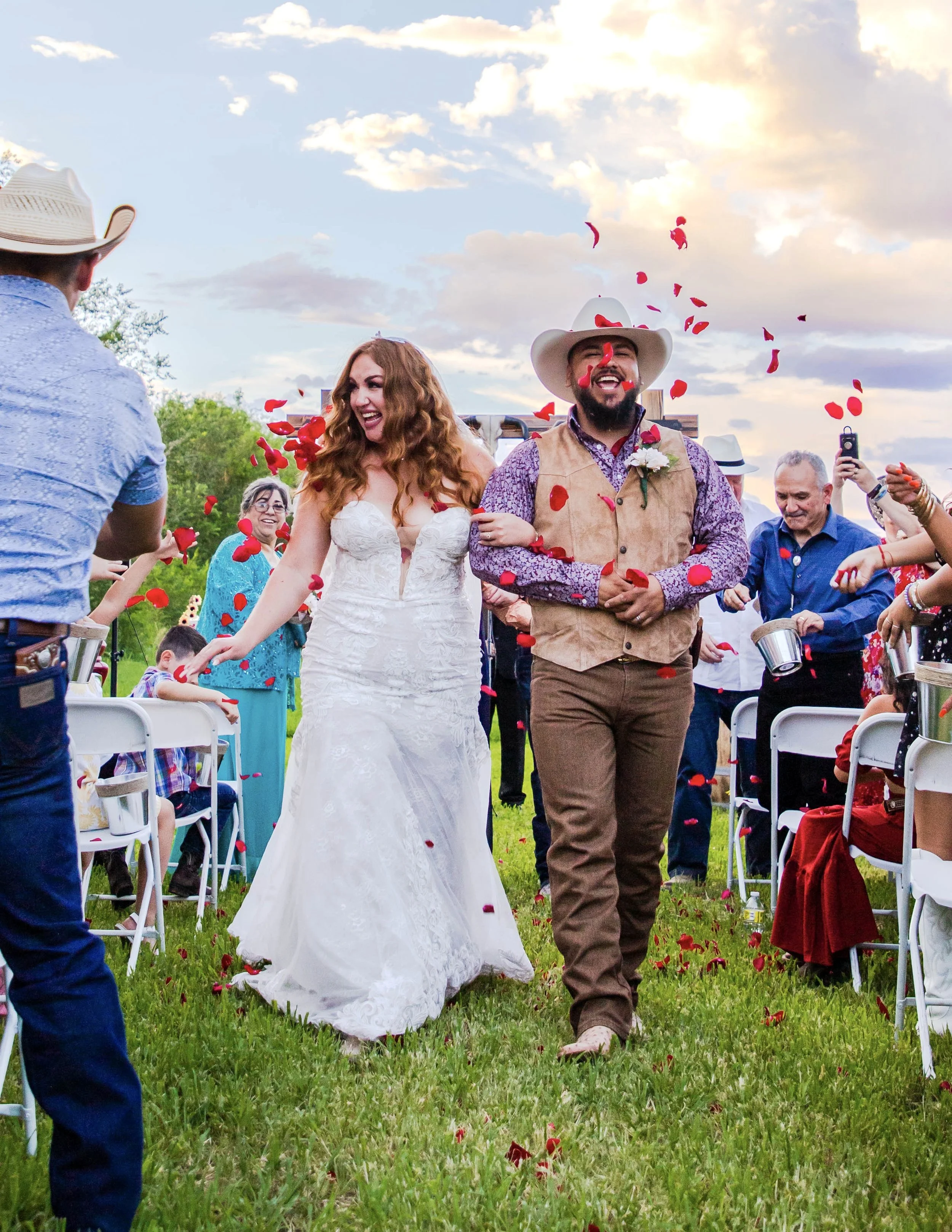 A bride and groom walk down the aisle at an outdoor wedding ceremony during sunset, celebrating with floating rose petals around them, surrounded by smiling guests.
