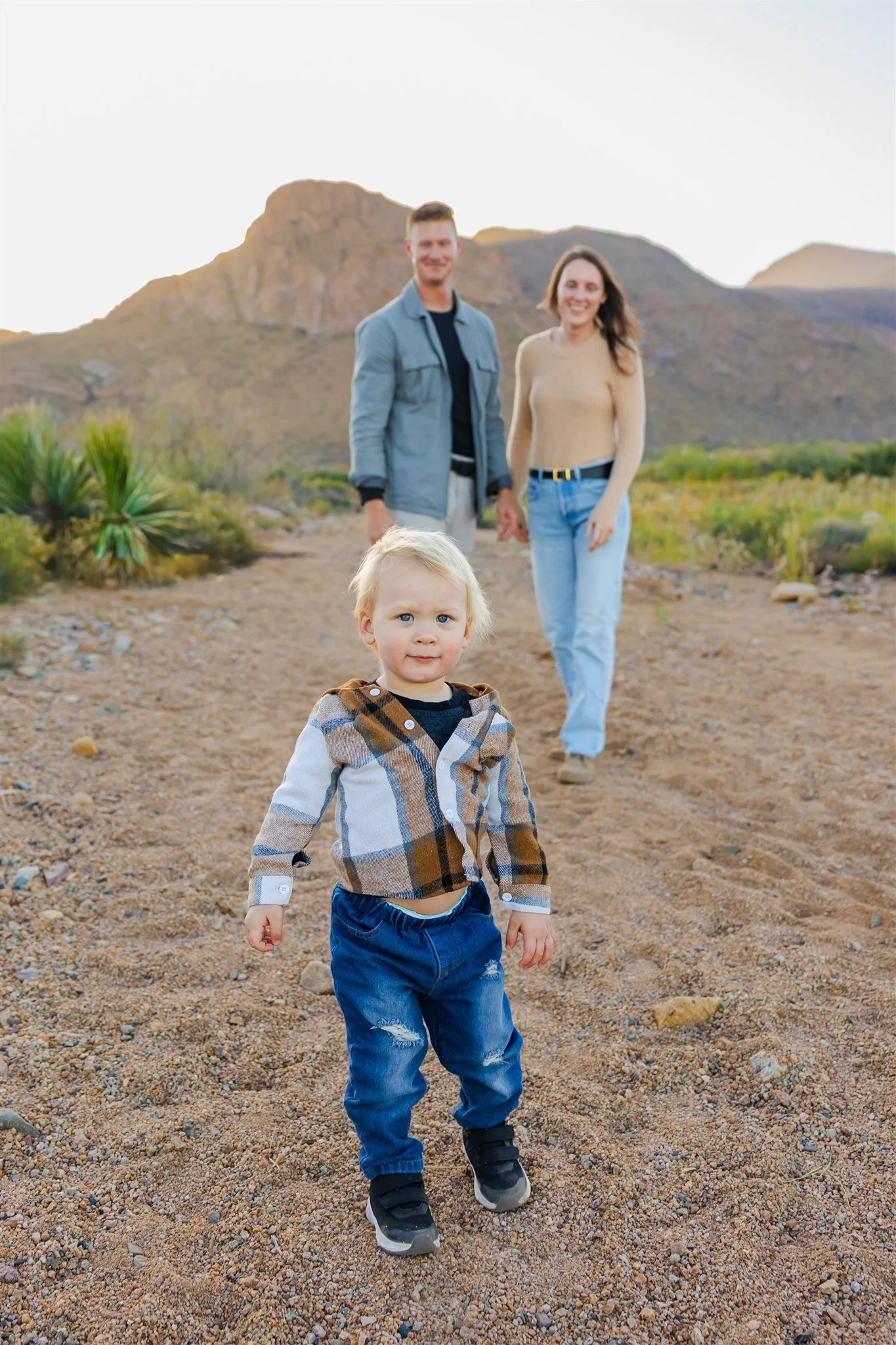 A young boy in a plaid jacket and blue jeans walking on a rocky trail with two smiling adults, a man and a woman, and a desert mountain landscape in the background.