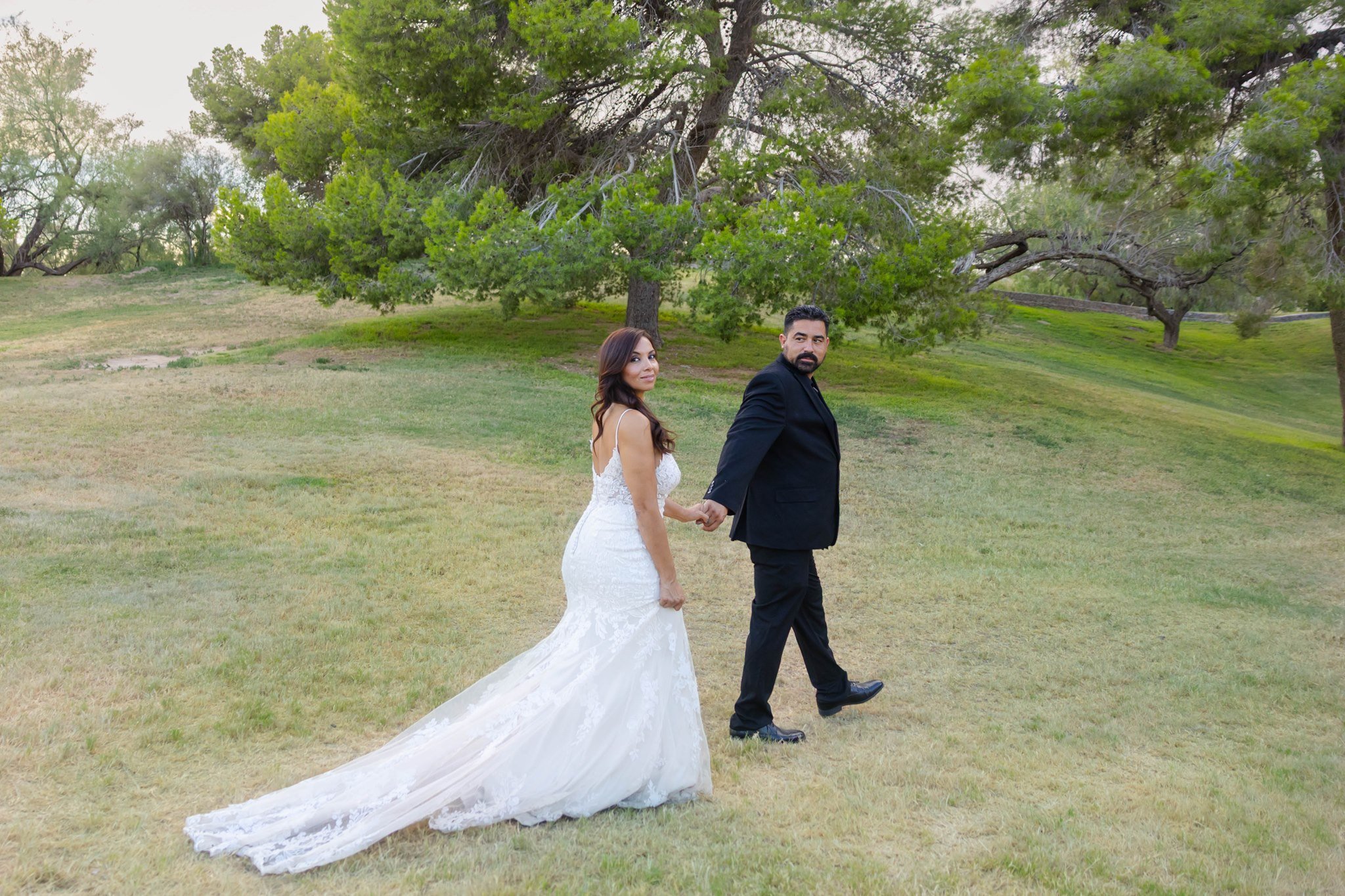 A newlywed couple holding hands and walking outdoors at Memorial park in El Paso, Texas with large green trees in the background. The bride is in a white wedding dress with a long train, and the groom is in a black suit.