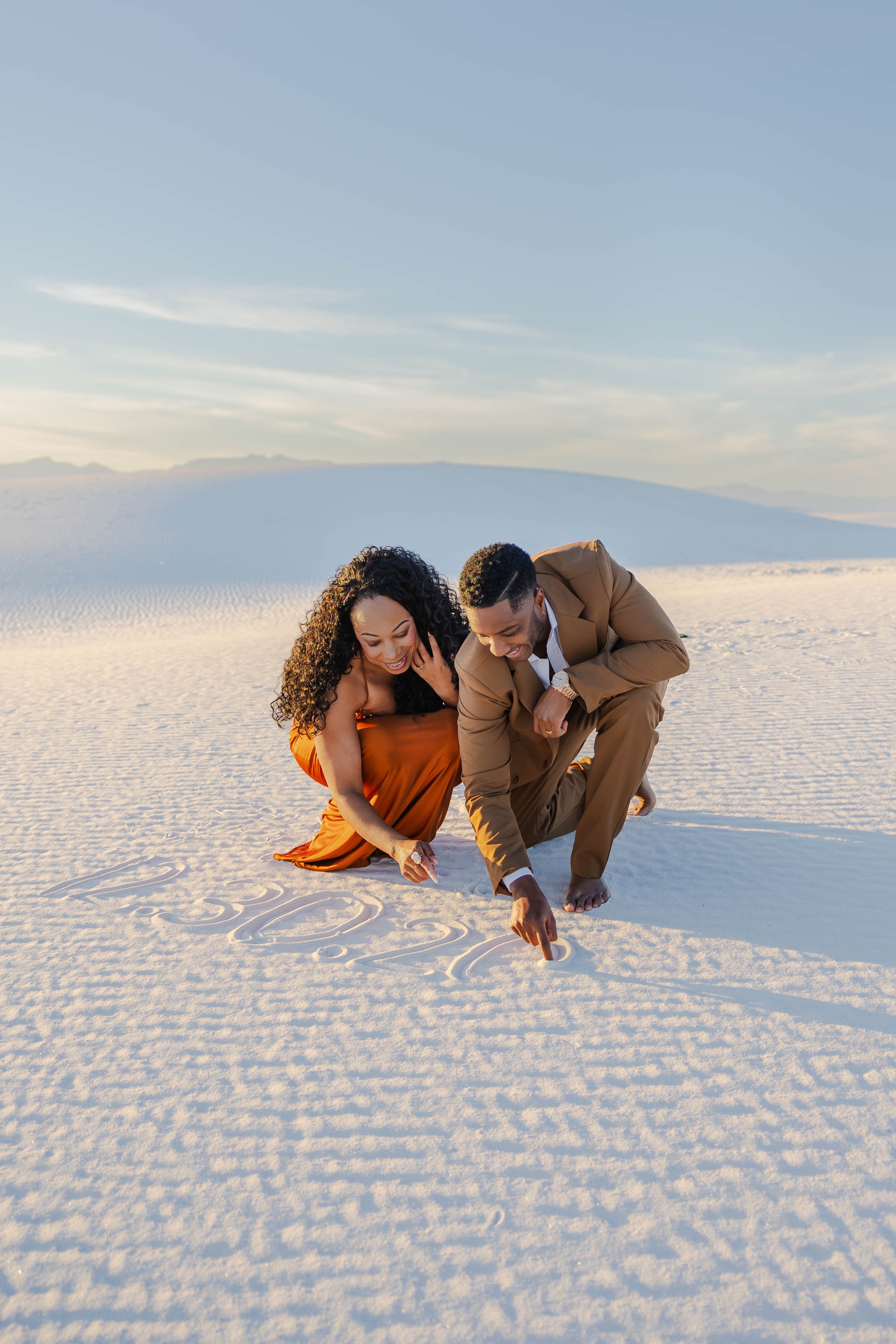 A man and woman crouching at White Sands National Park, drawing in the sand with their fingers with a mountain background and blue sky.