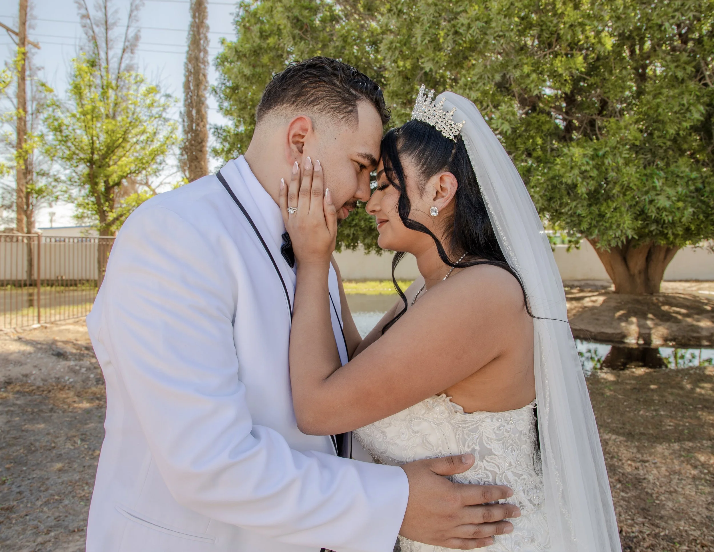 A bride and groom at Socorro Mission Church touching foreheads, outdoors with trees in the background.
