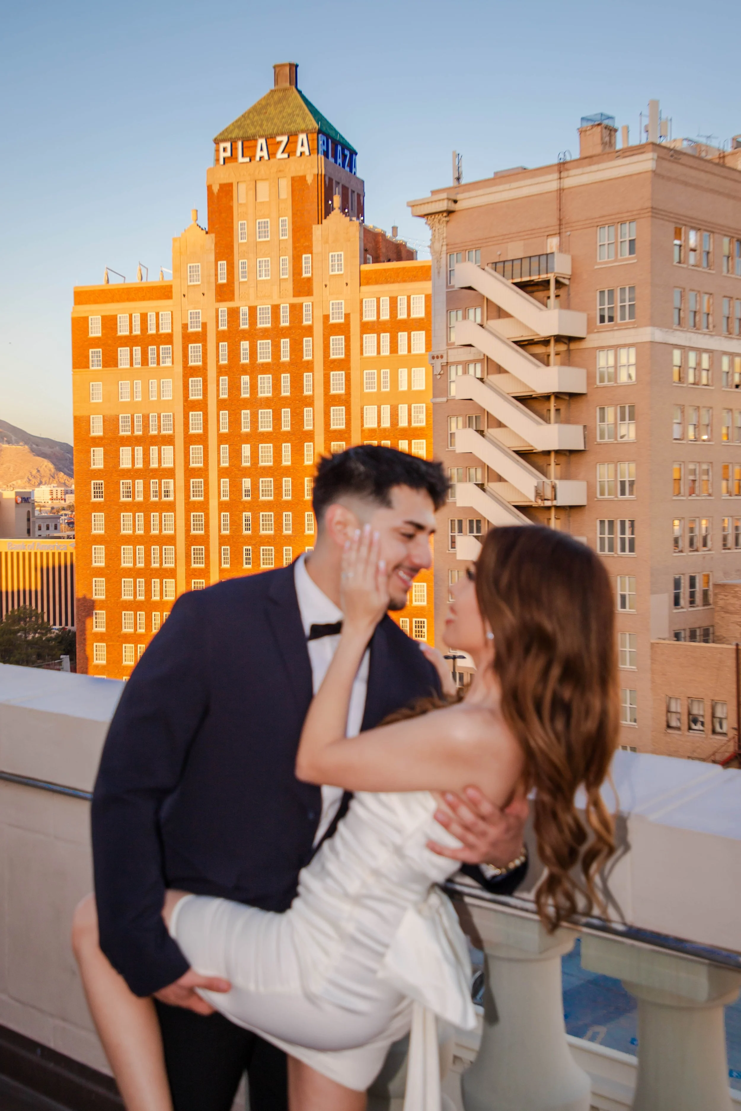 An engaged El Paso couple dressed in formal attire sharing a romantic moment on a rooftop balcony with The Plaza Theatre in the background, during sunset.