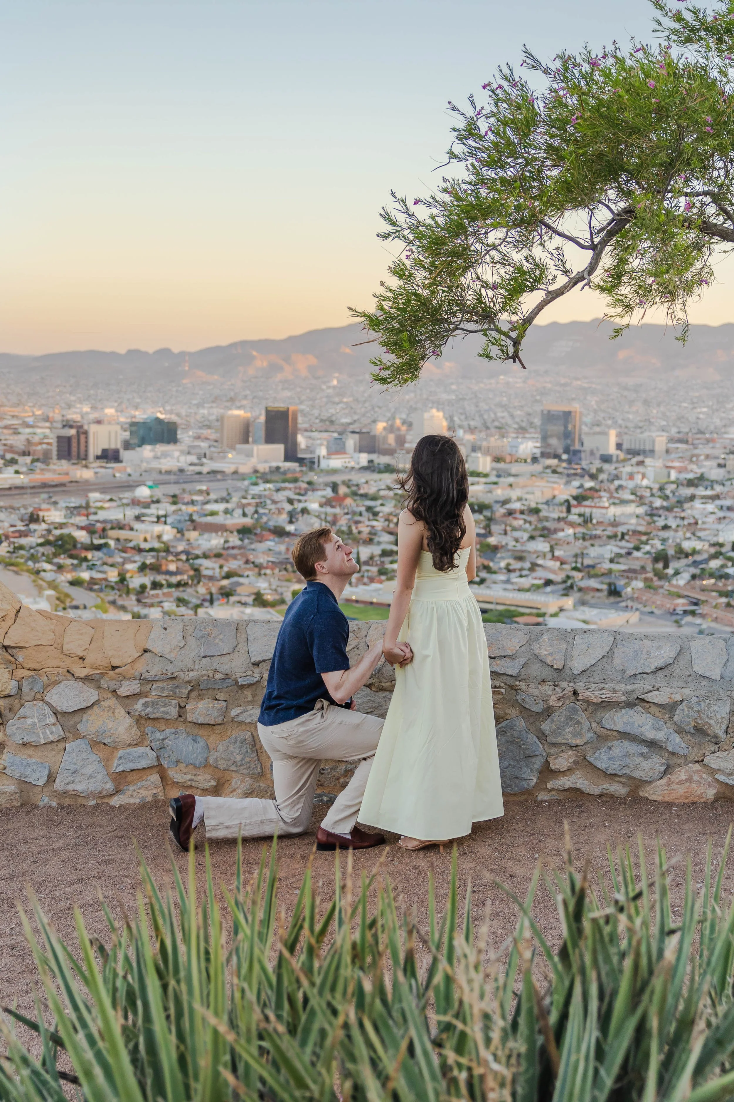 A man is proposing marriage to a woman outdoors at El Paso's Scenic Drive during sunset. The man is kneeling, holding the woman's hand, while the woman is standing and looking down at him.