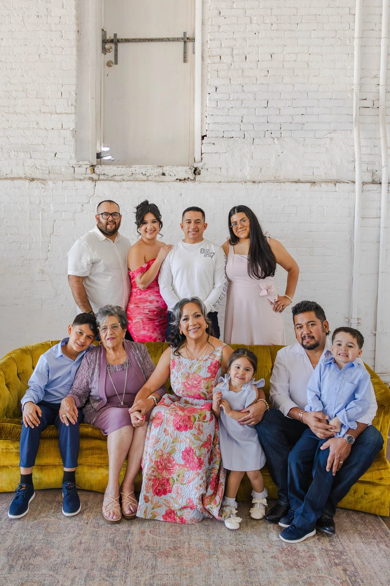 Family group photo in a modern studio with a white brick wall, including three generations: elderly woman, middle-aged adults, and young children seated on a yellow velvet sofa.