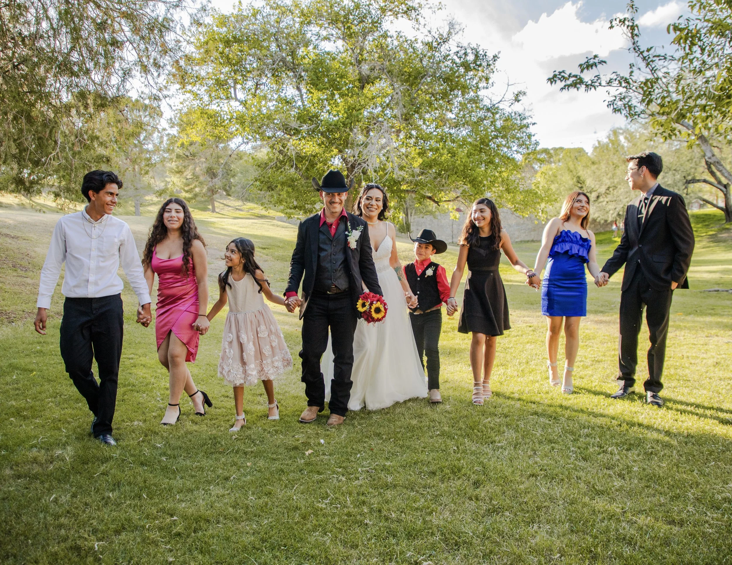 Family holding hands and smiling, walking at Memorial Park on the grass during daytime, with trees and blue sky in the background, celebrating a wedding.