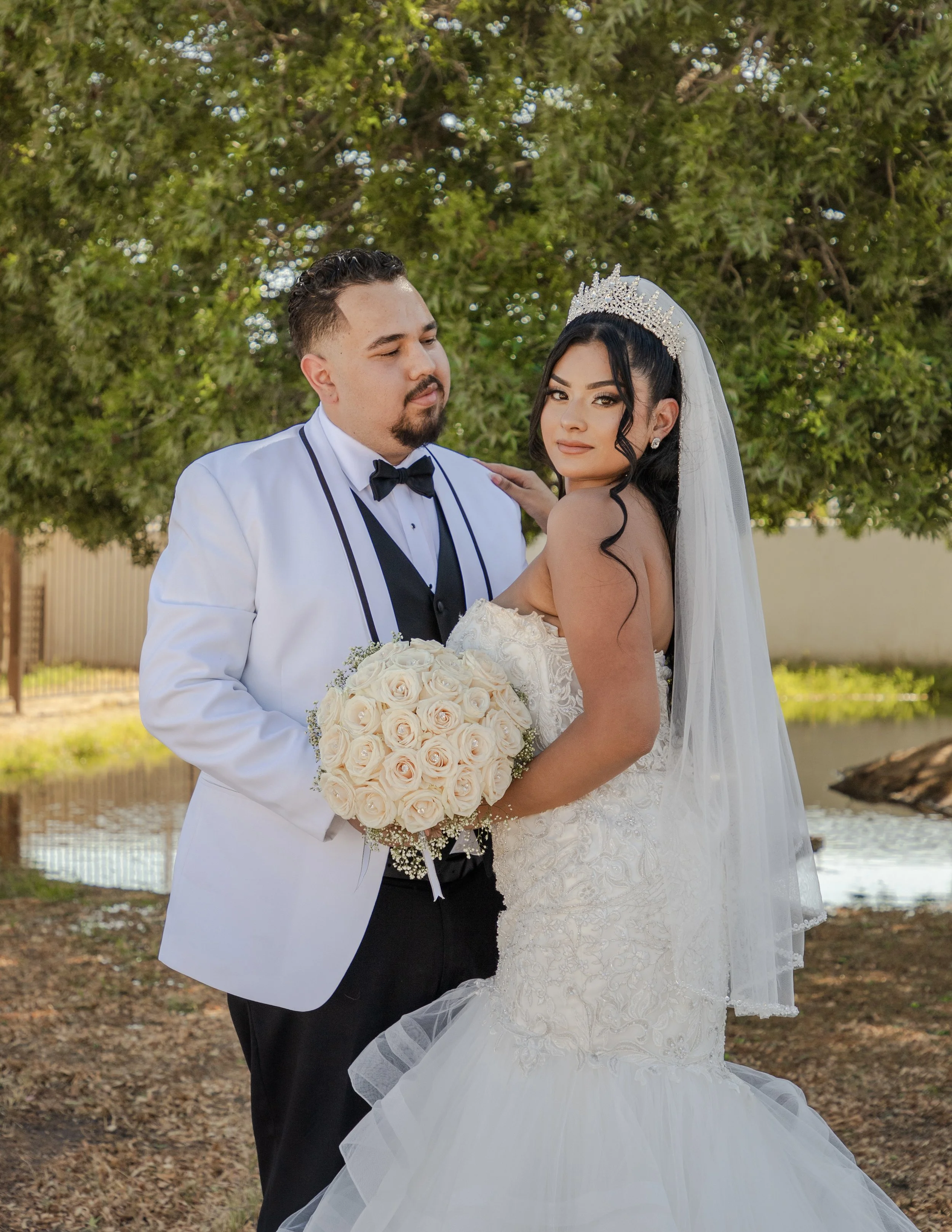 A bride and groom pose outdoors in front of a large green tree at the Socorro Mission Church in El Paso, with the bride holding a bouquet of white roses.