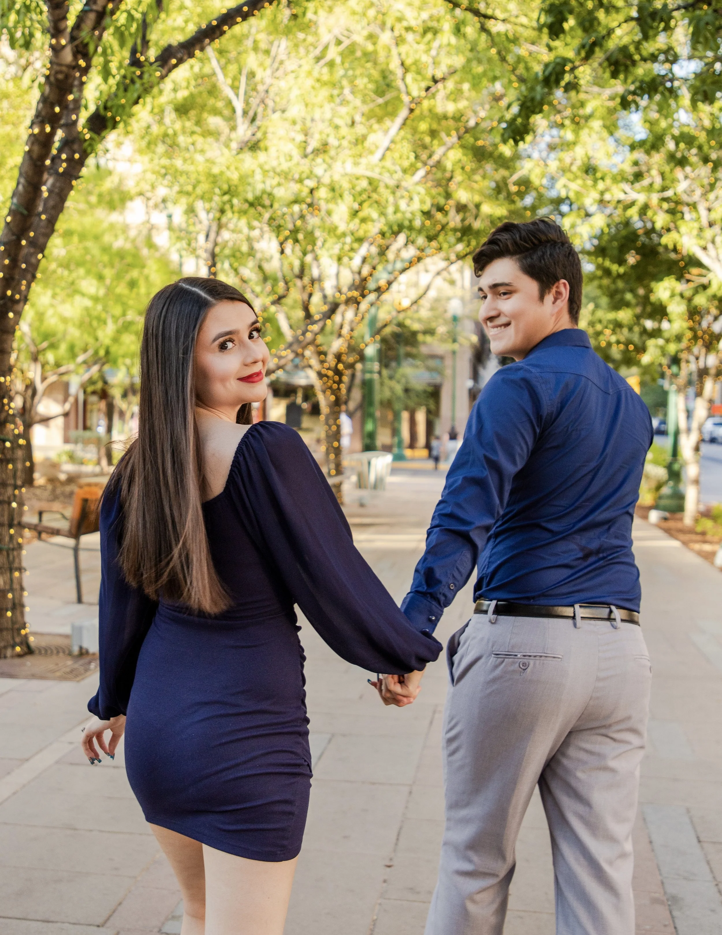 A couple holding hands and looking at each other in Downtown El Paso during the daytime, with trees decorated with string lights in the background.