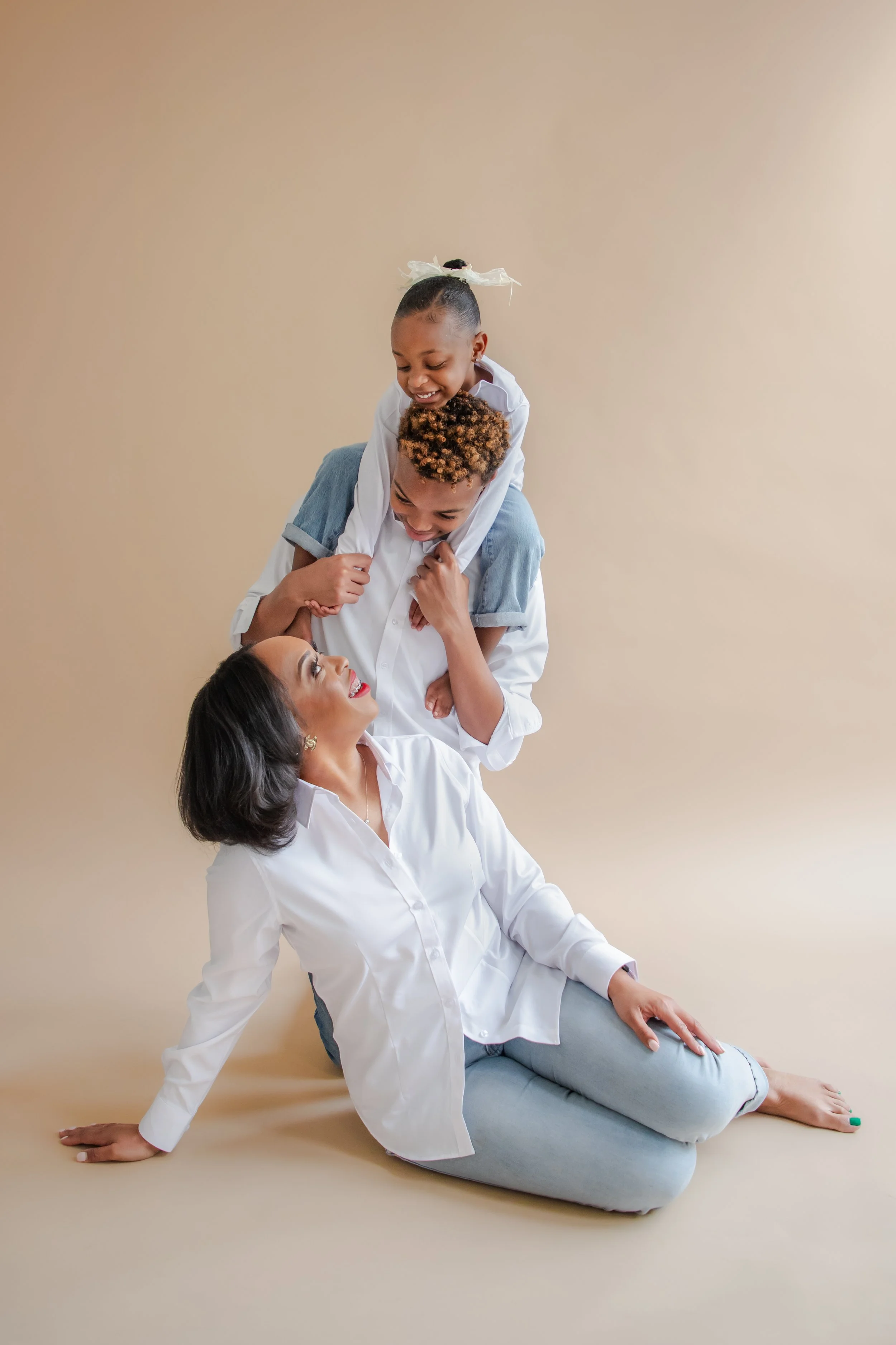 A woman and two children playing together, with one child sitting on the woman's shoulders and the other child sitting on top of the first child. They are smiling and looking at each other, against a plain beige background.