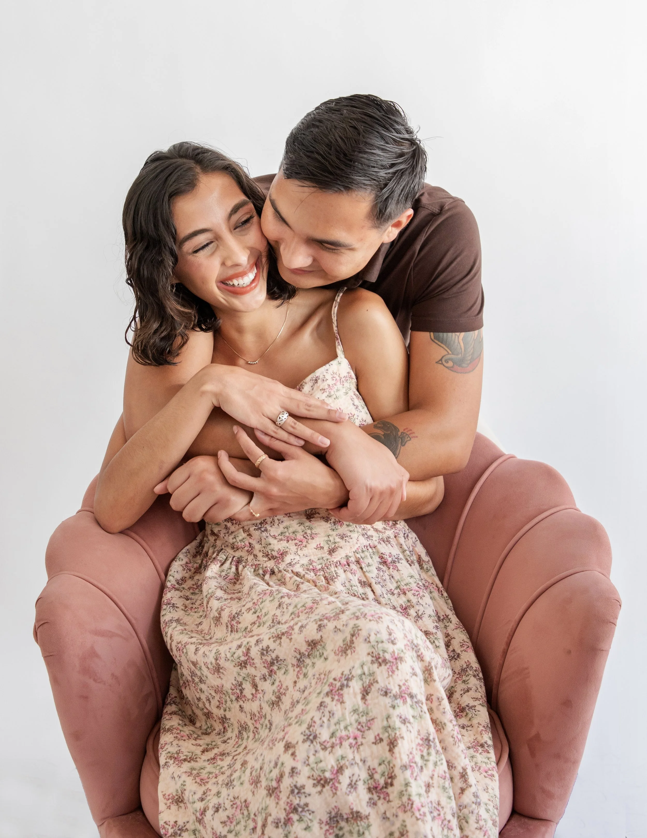 A woman with dark hair and a man with dark, slicked-back hair smile and embrace as they sit on a pink velvet armchair against a plain white background.