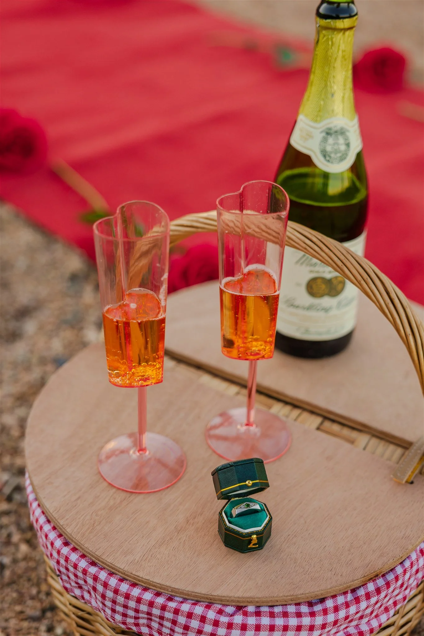 A small round wooden table with a red tablecloth, set with two tall champagne glasses filled with a pink drink, a bottle of sparkling wine in the background, a small ring box, and a black jewelry box, at El Paso's Franklin Mountains.