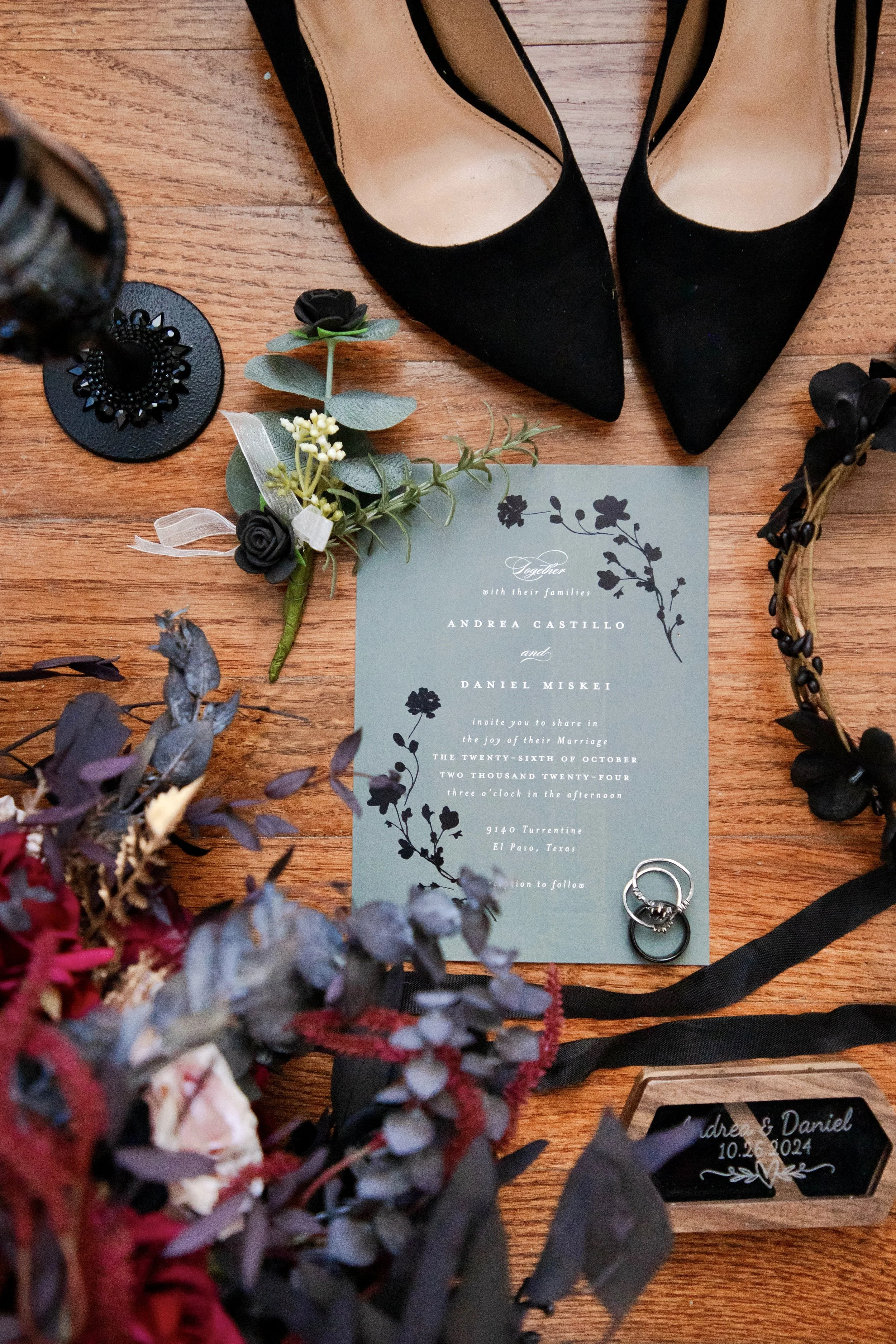 A pair of black high-heeled shoes, a wedding invitation, some jewelry including rings and a bracelet, and floral decorations are arranged on a wooden surface.