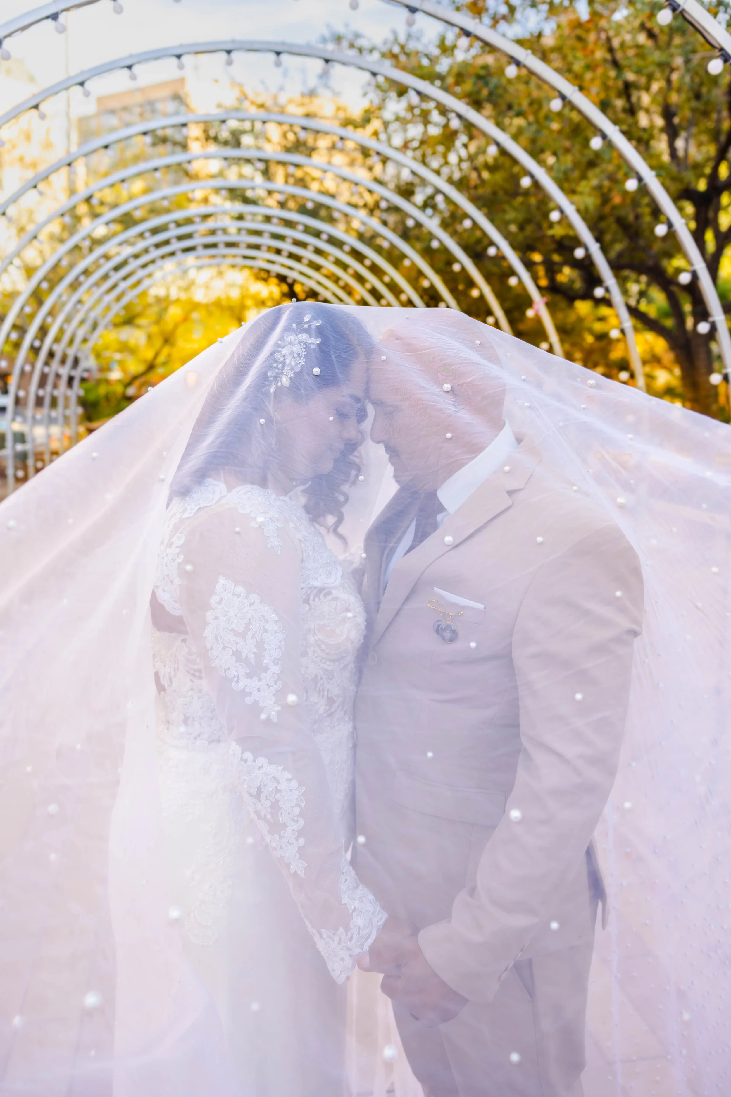 A bride and groom face to face under a sheer veil that covers both their faces. The background features autumn trees and a series of white arches with lights in Downtown El Paso during winter.