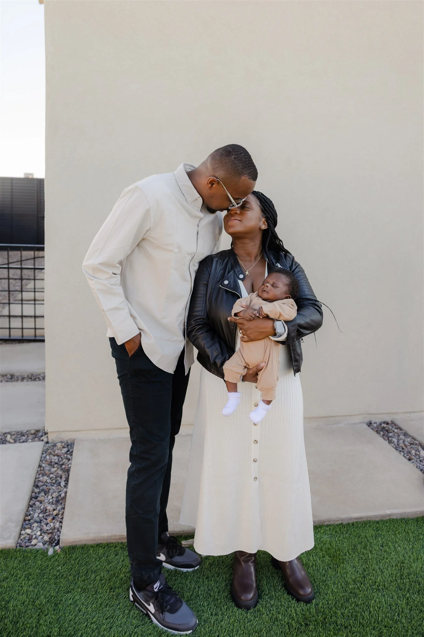 A woman holding a sleeping baby with a man leaning in to kiss her forehead. They are standing outdoors on a small patch of artificial grass with a plain wall behind them.