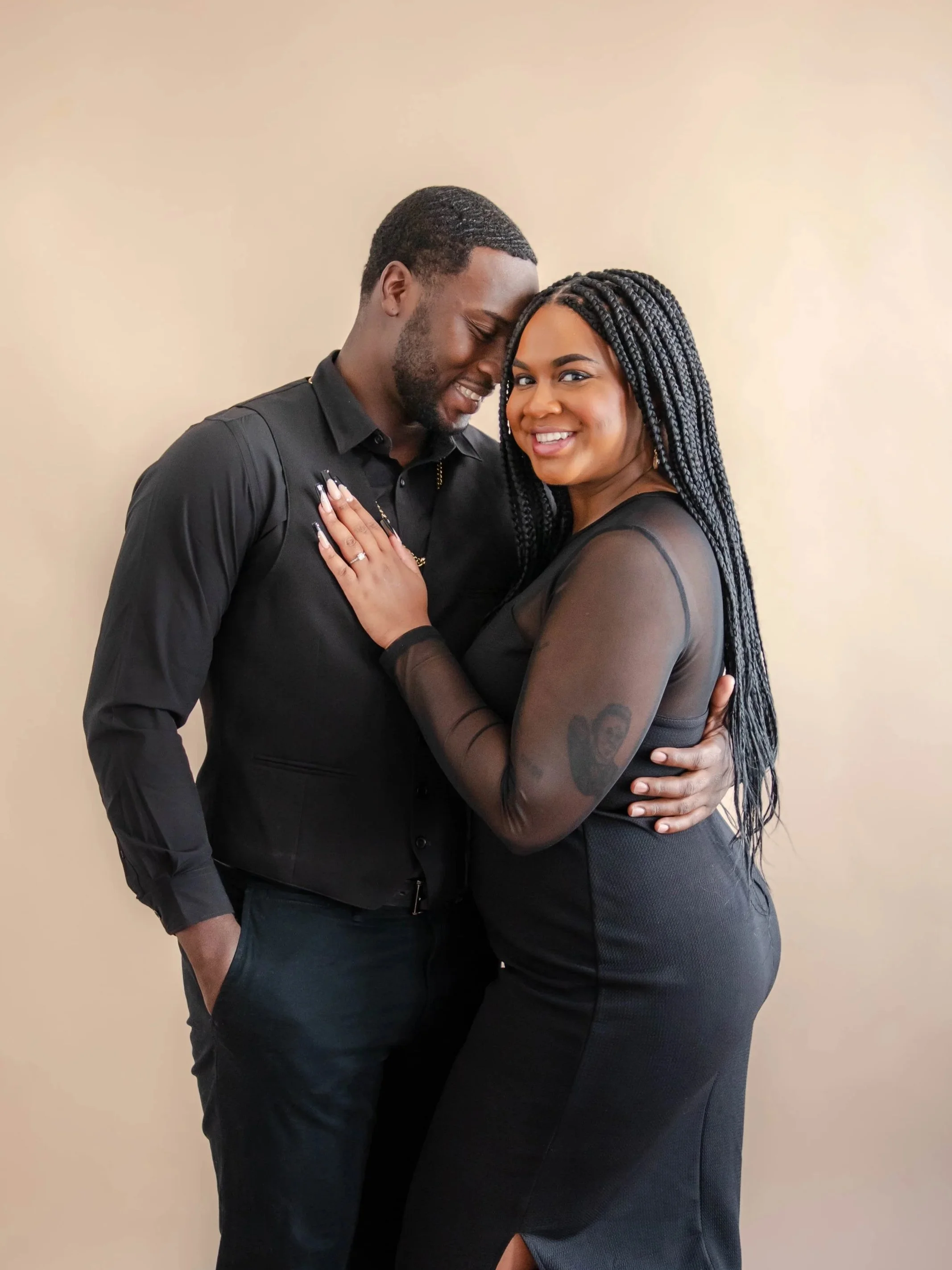 An El Paso couple standing close together against a plain beige wall, smiling and embracing each other. The woman has long braided hair and is wearing a black sheer top with a skirt. This is at The Brewhouse Studio.