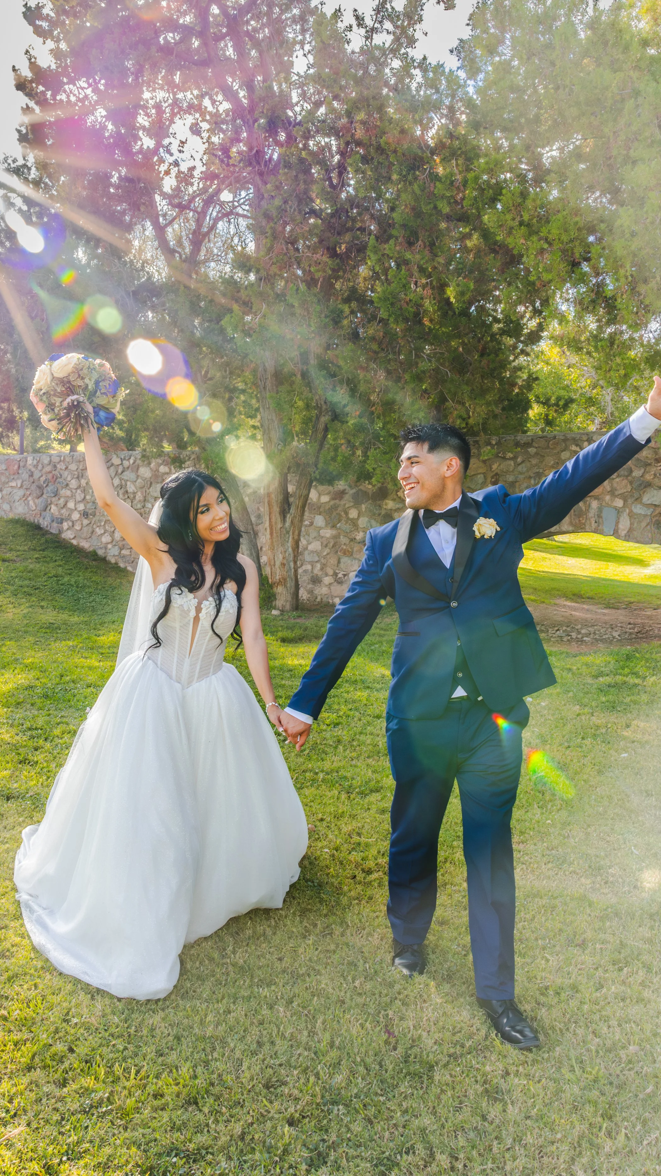 A joyful bride and groom holding hands outdoors on a sunny day. The bride is wearing a white wedding dress and holding a bouquet, while the groom is dressed in a dark blue suit with a black bow tie. They are smiling and celebrating, with large trees and a stone wall in the background, and sunlight creating lens flares in the image.