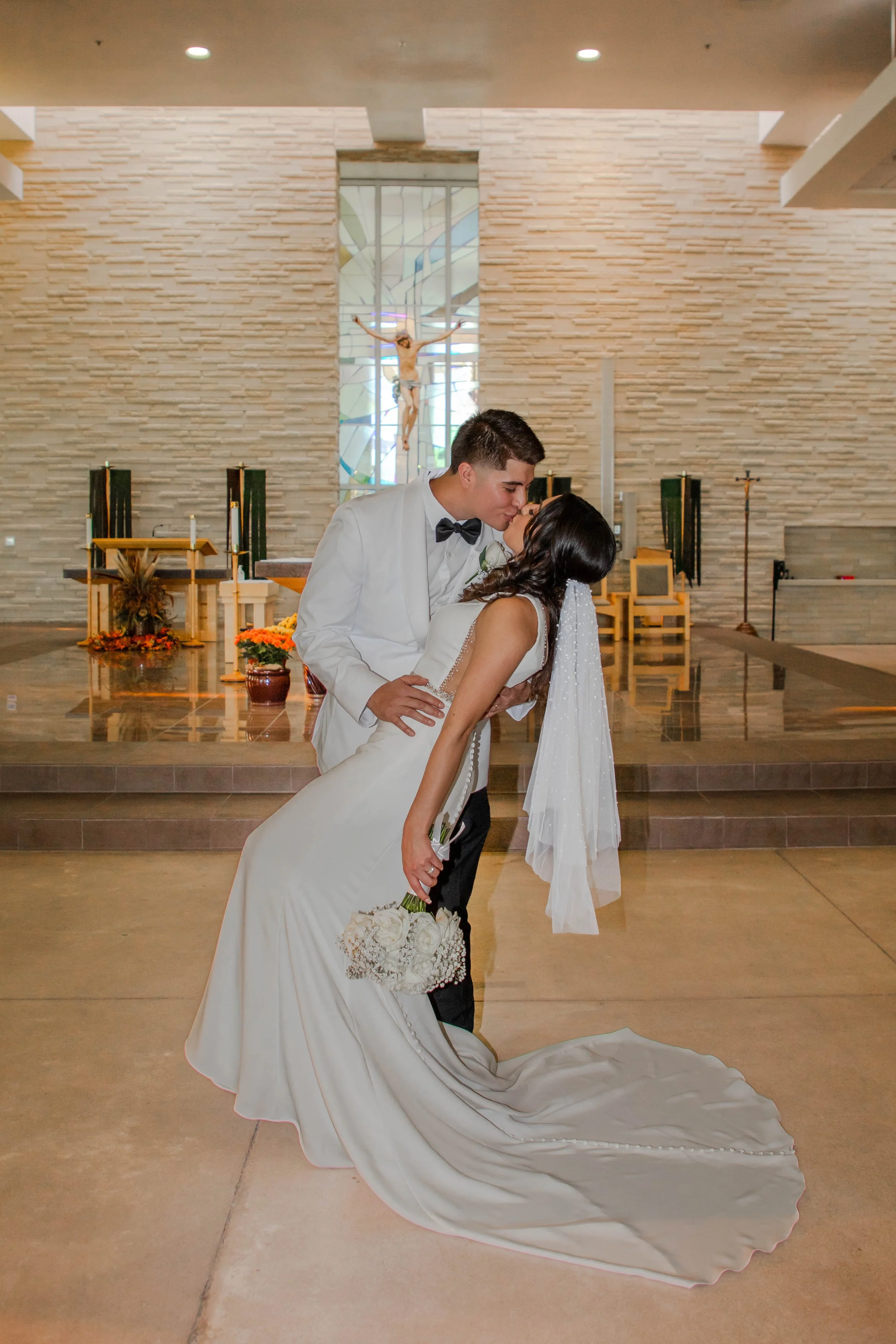 A bride and groom sharing a kiss at St. Stephens church after their wedding, with a stained glass window and crucifix in the background.