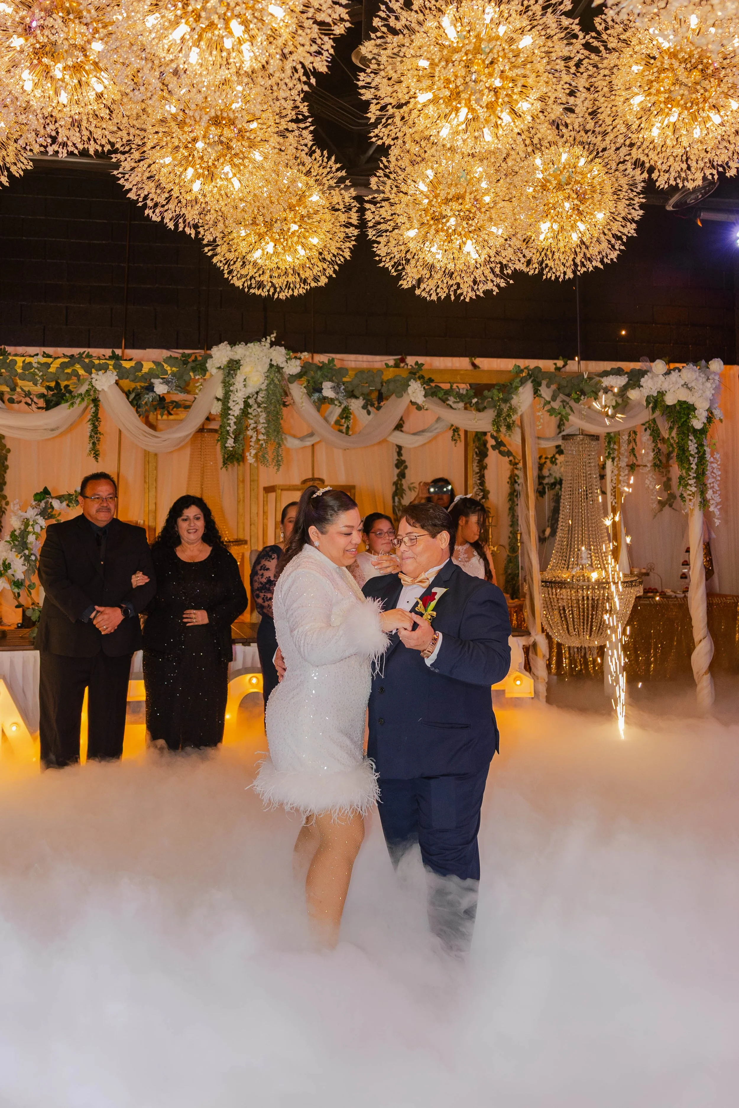 A married couple dancing at their wedding reception in El Paso, Texas surrounded by guests, with elaborate golden chandeliers and floral decorations overhead, and fog on the floor.