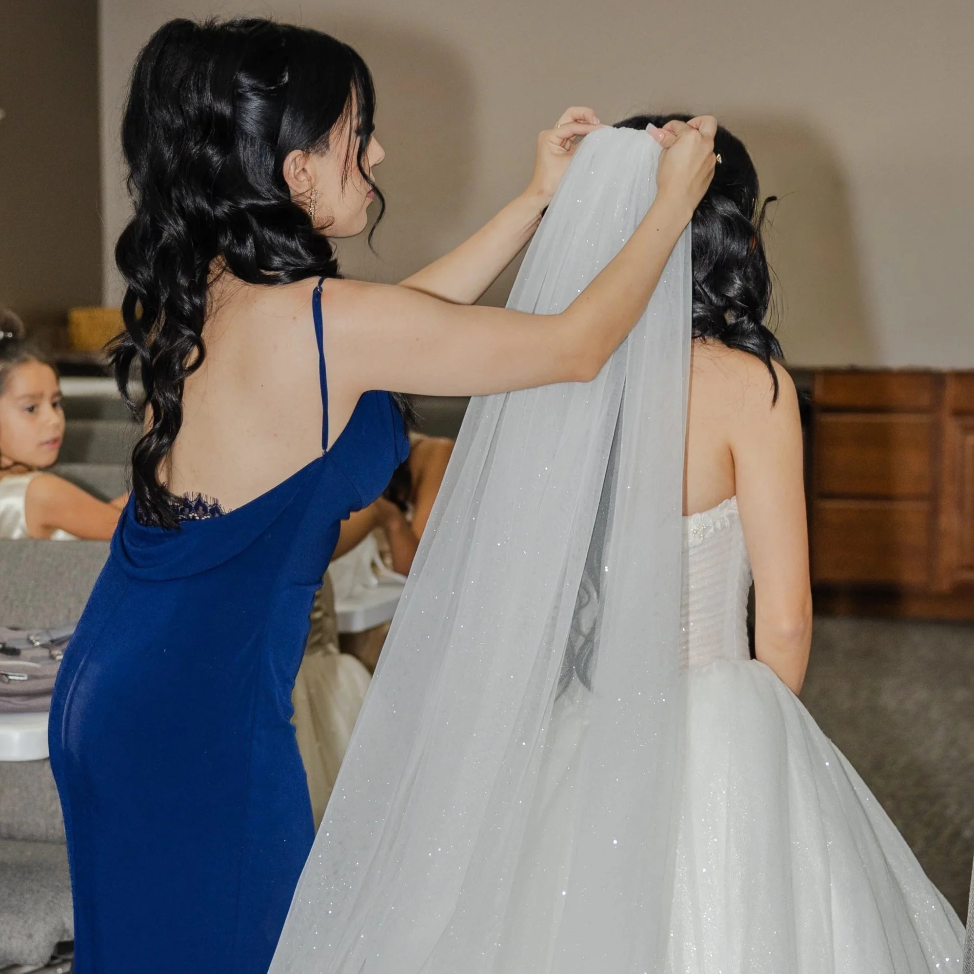 A woman with black, wavy hair, wearing a blue dress, helping a bride put on a white, sparkly tulle veil at a wedding in El Paso.