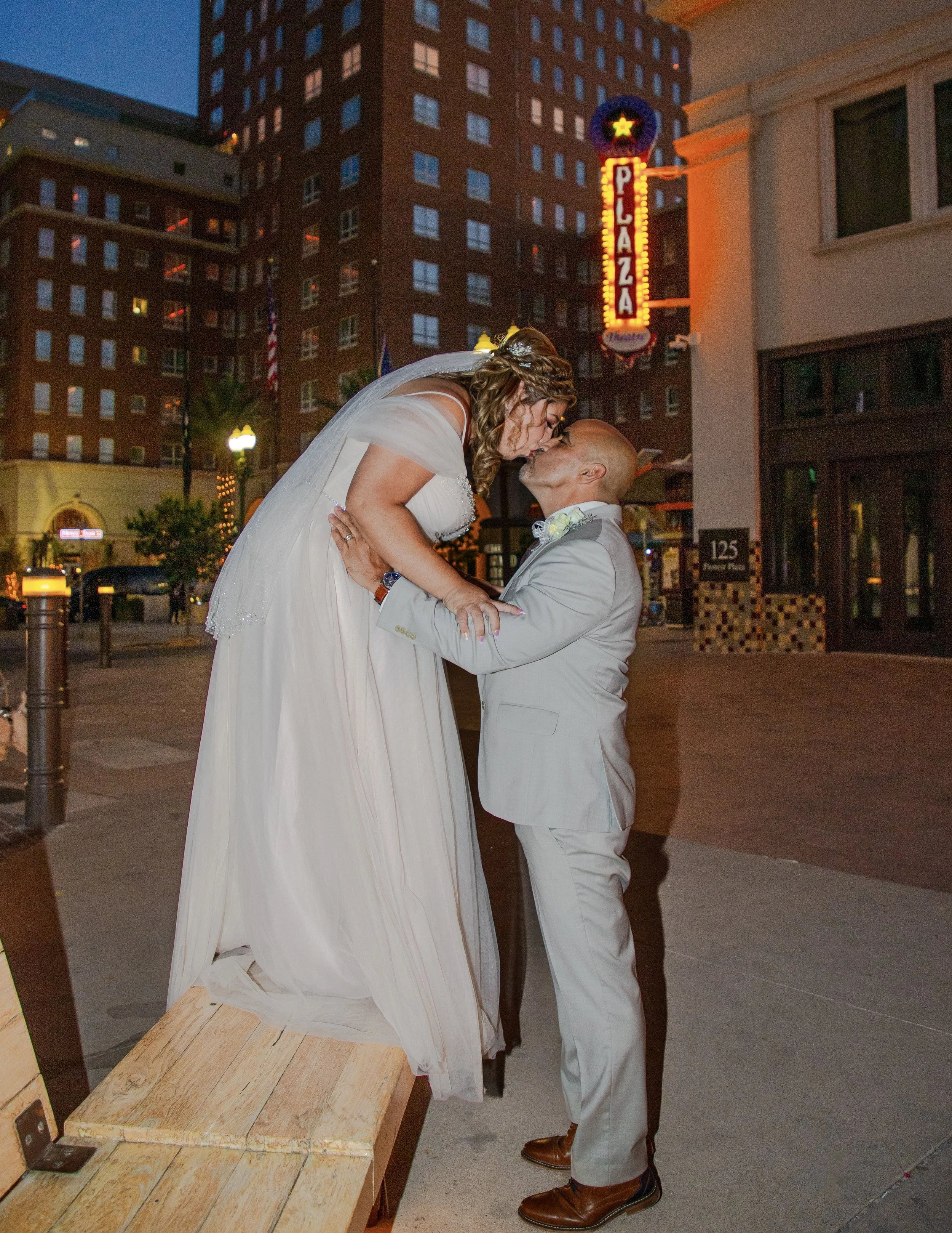A newlywed couple sharing a kiss on a city street at dusk, with a neon pizza sign and buildings in the background.