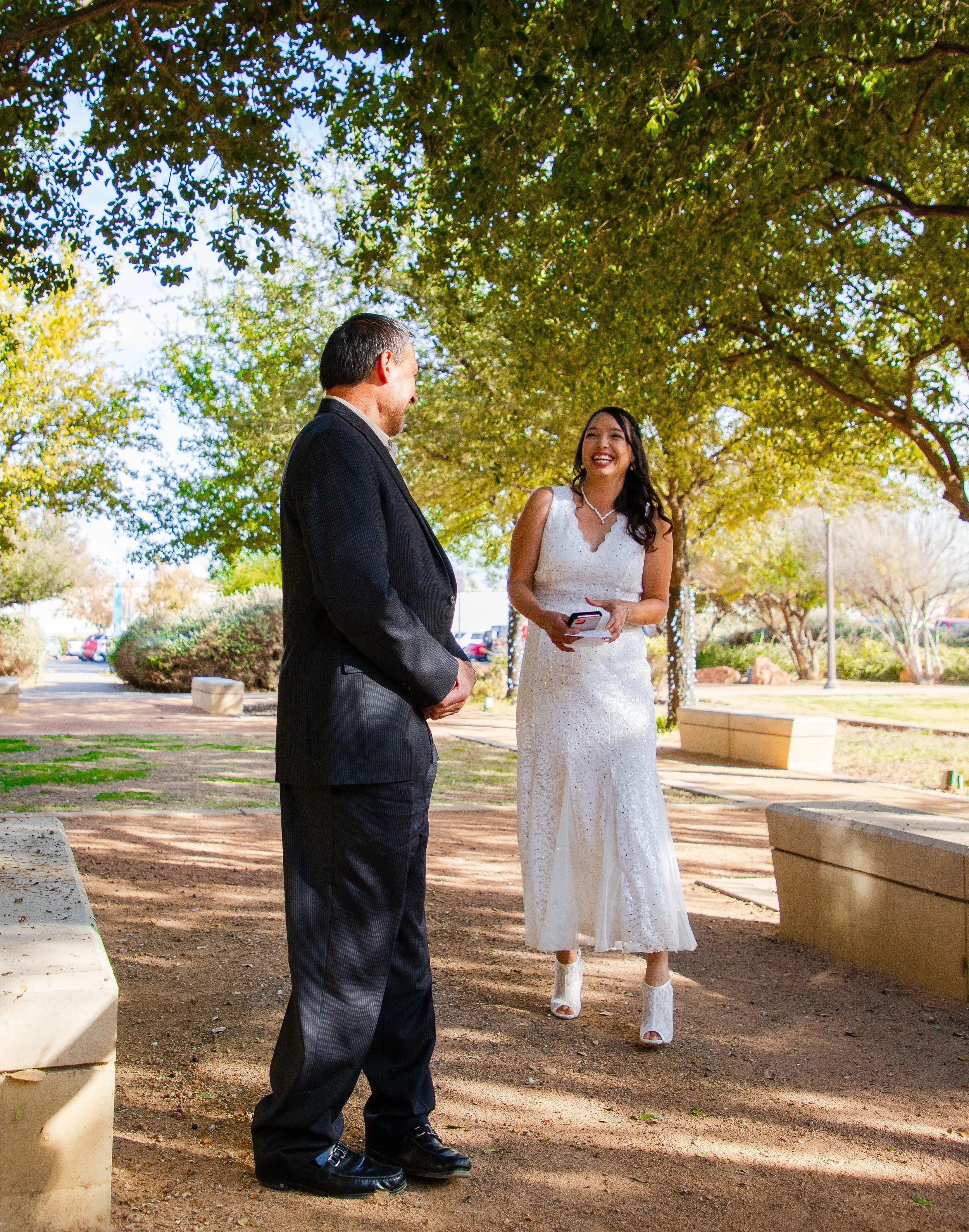 A man in a suit and a woman in a white dress are standing at El Paso's Texas Tech campus under trees with sunlight filtering through the leaves. They are smiling and appear to be having a conversation.