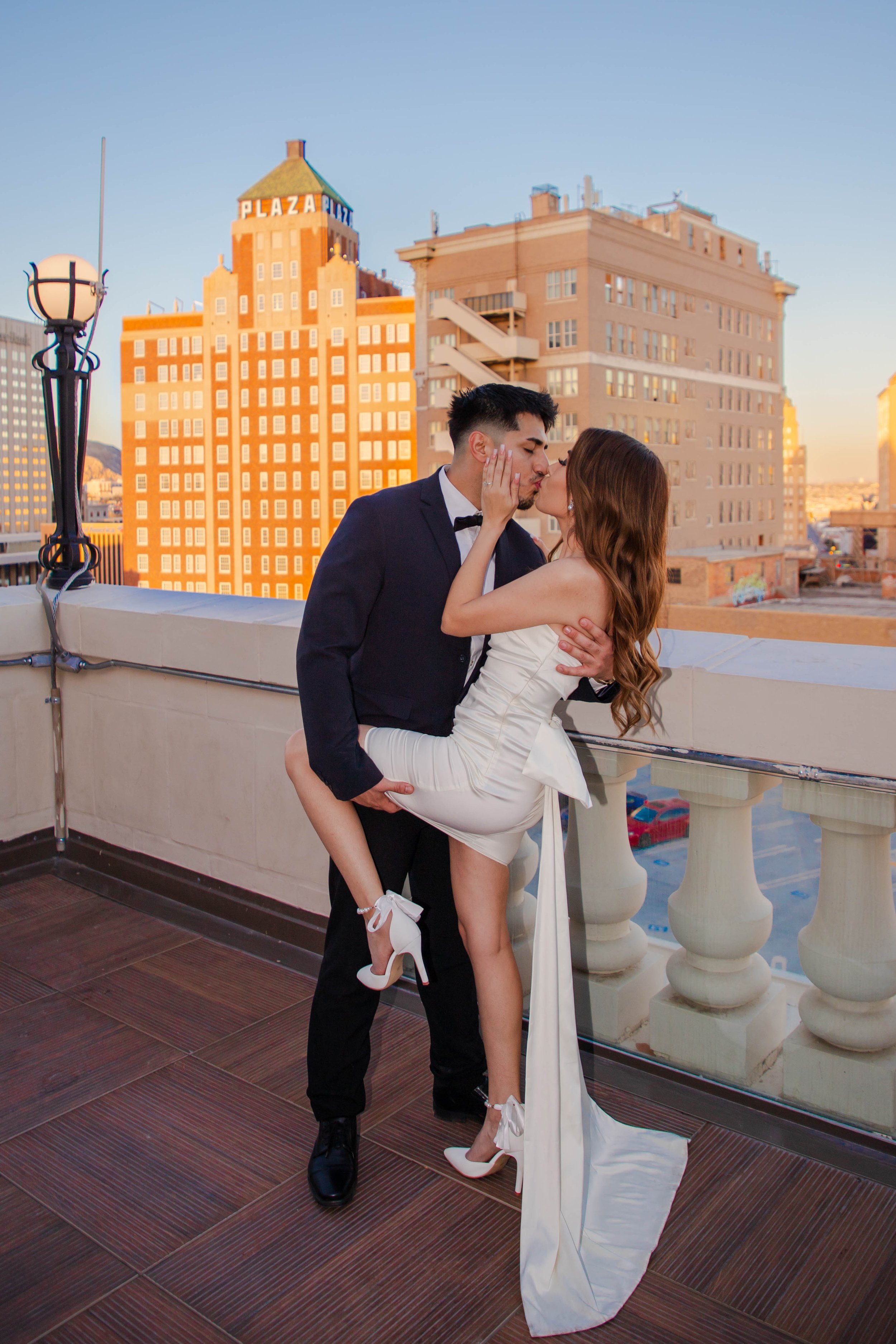 An engaged couple kissing on an El Paso rooftop deck at sunset, with the Plaza Theatre and buildings in the background. The woman and man are doing a dip kiss.