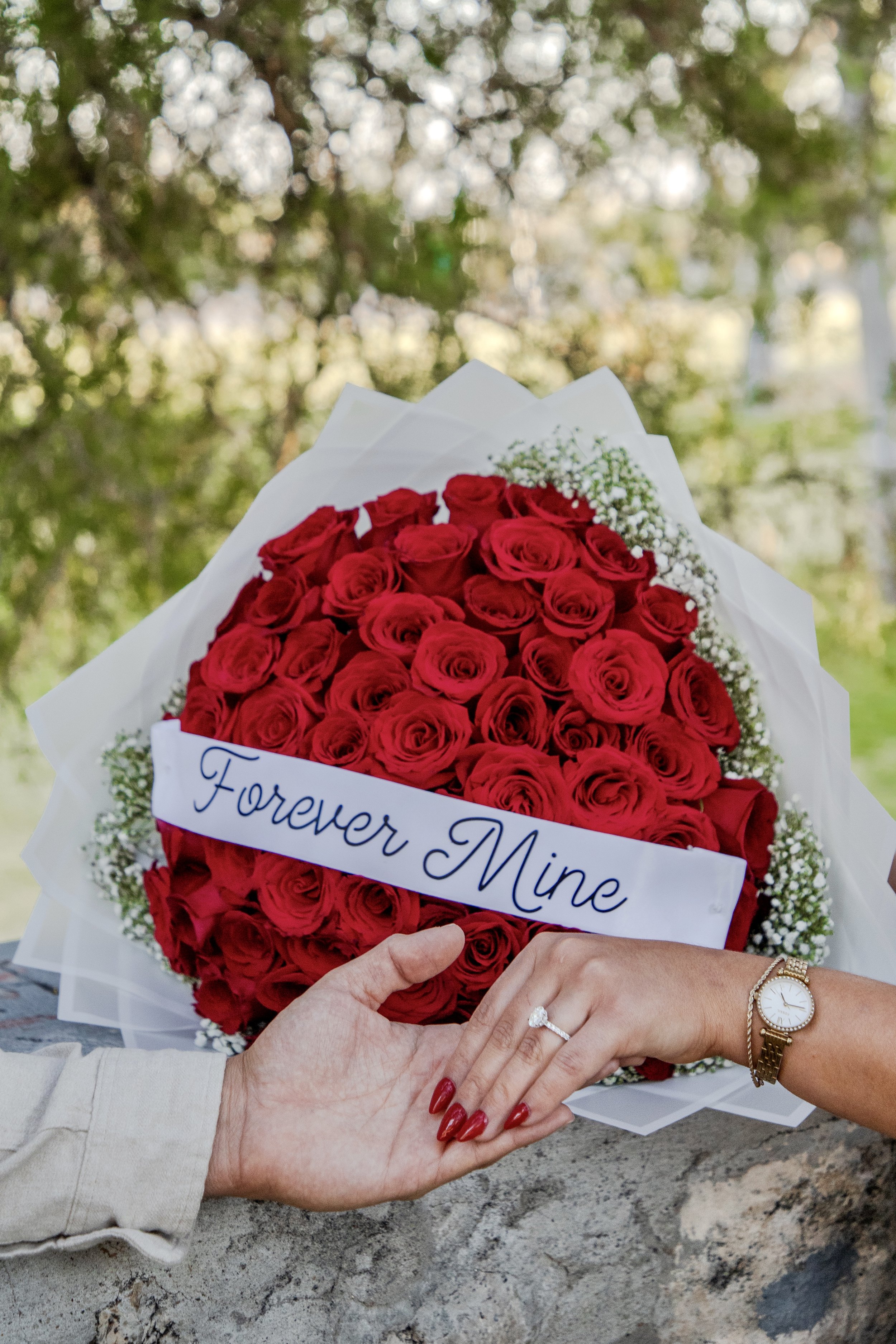 A bouquet of red roses wrapped in white paper with a banner saying "Forever Mine" is held by two women, one with red nail polish and a watch, and the other with a diamond ring and a bracelet, in an outdoor setting with blurred trees in the background.