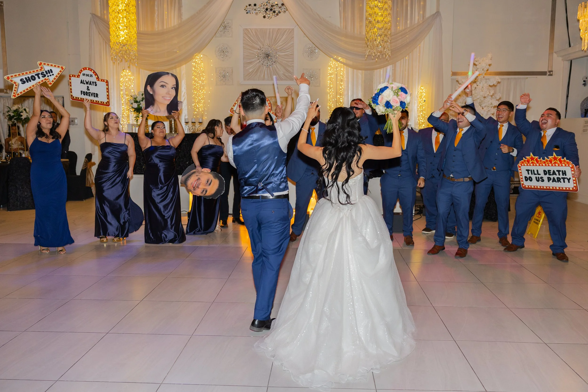 A wedding reception with a bride and groom dancing among friends and family at Bebe Hall in El Paso. Guests are holding signs with messages and portraits, and there are decorative lights and drapes in the background.