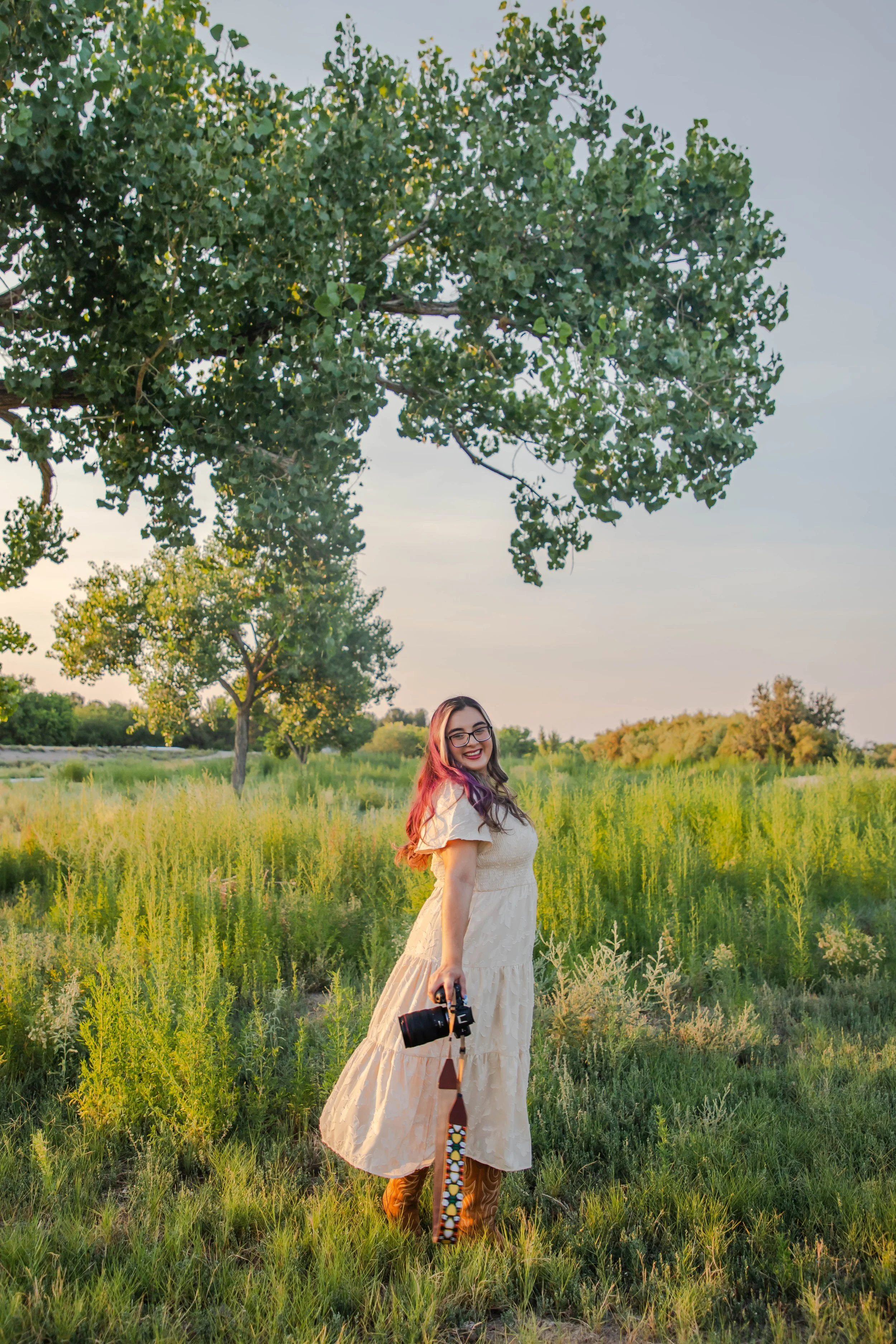 A woman with glasses, long hair with pink streaks, and a beige dress, holding a camera, standing in a green field near trees during sunset.