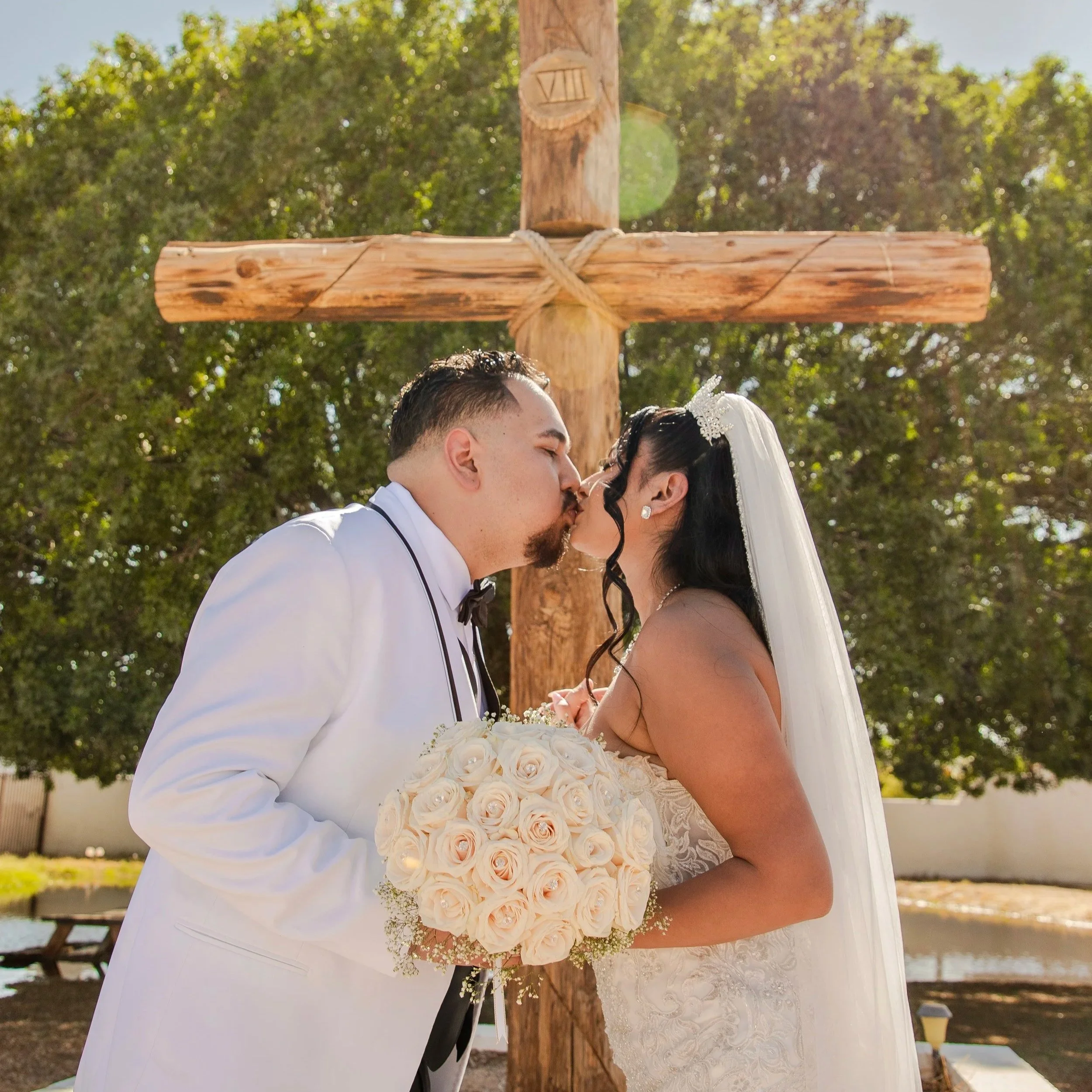 A couple kissing at their wedding in the Socorro Mission, the bride holding a bouquet of white roses, standing outdoors with trees and a wooden cross in the background.