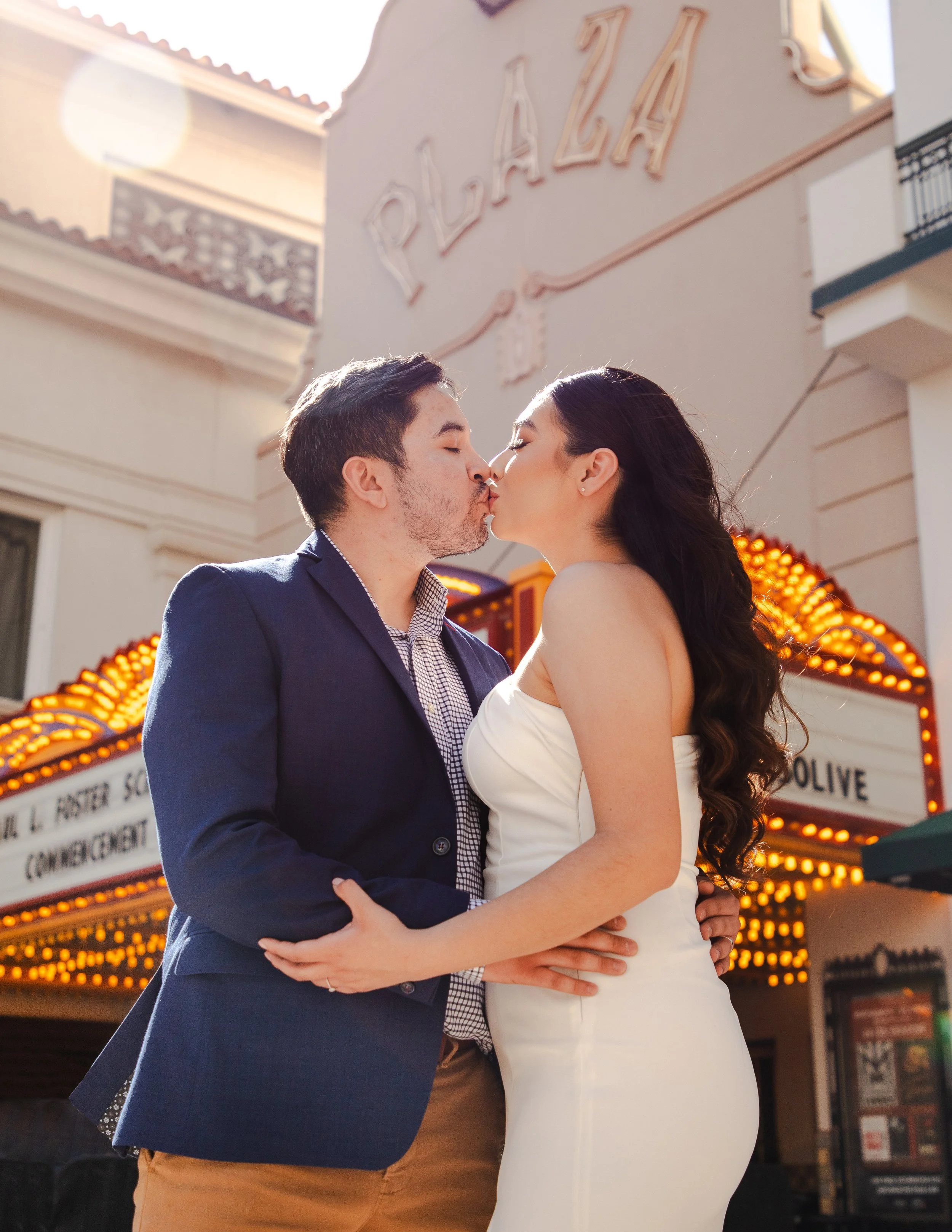 An eloped couple kissing in front of El Paso's Plaza Theatre during the evening. The man is wearing a navy blazer, and the woman is in a white wedding dress.