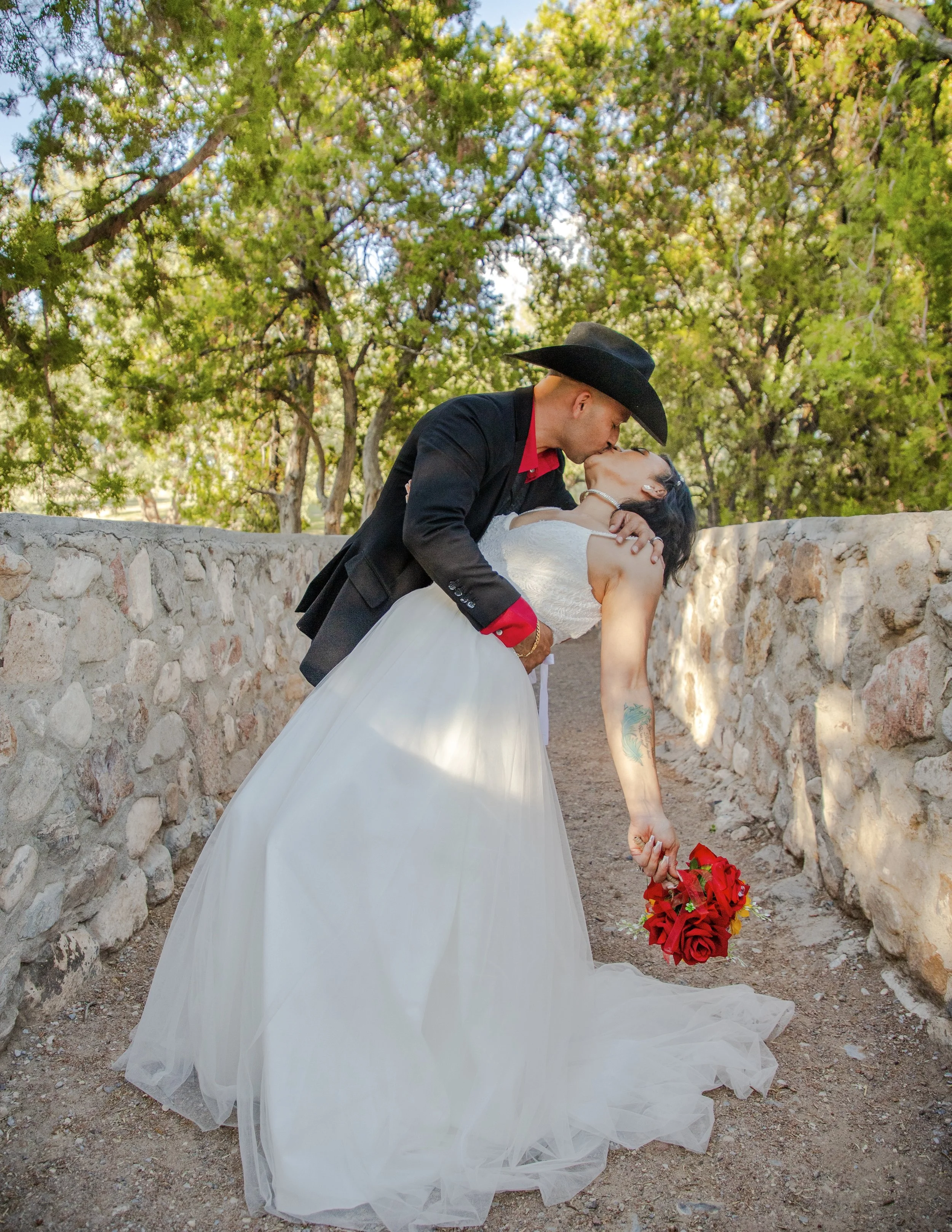 A man in a black cowboy hat and dark suit is dipping a woman in a white wedding dress in a romantic kiss outdoors. The woman holds a bouquet of red roses, and they are surrounded by green trees and a stone wall.