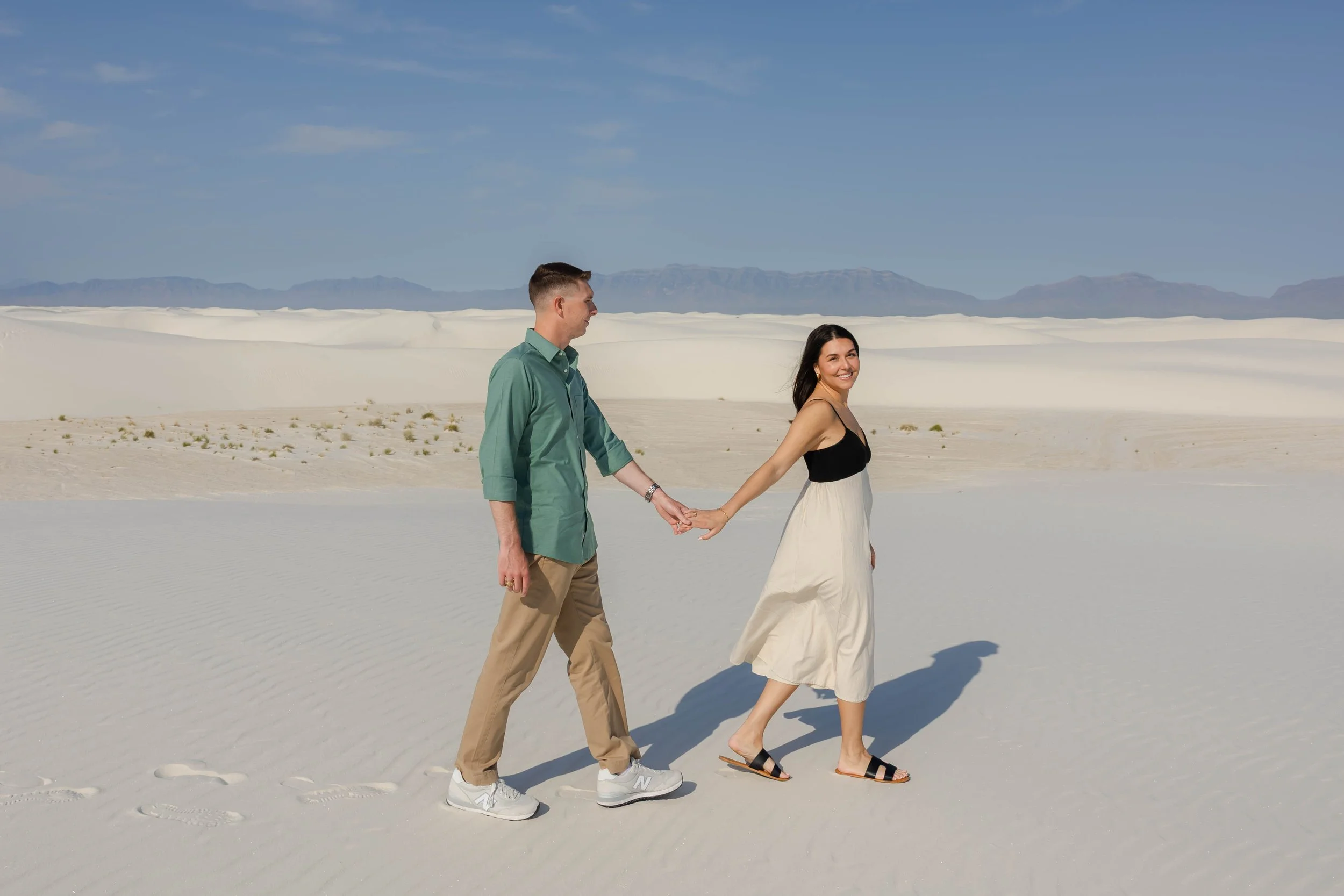 A couple walking hand-in-hand at White Sands National Park in New Mexico with a clear blue sky and the Organ mountains in the background.