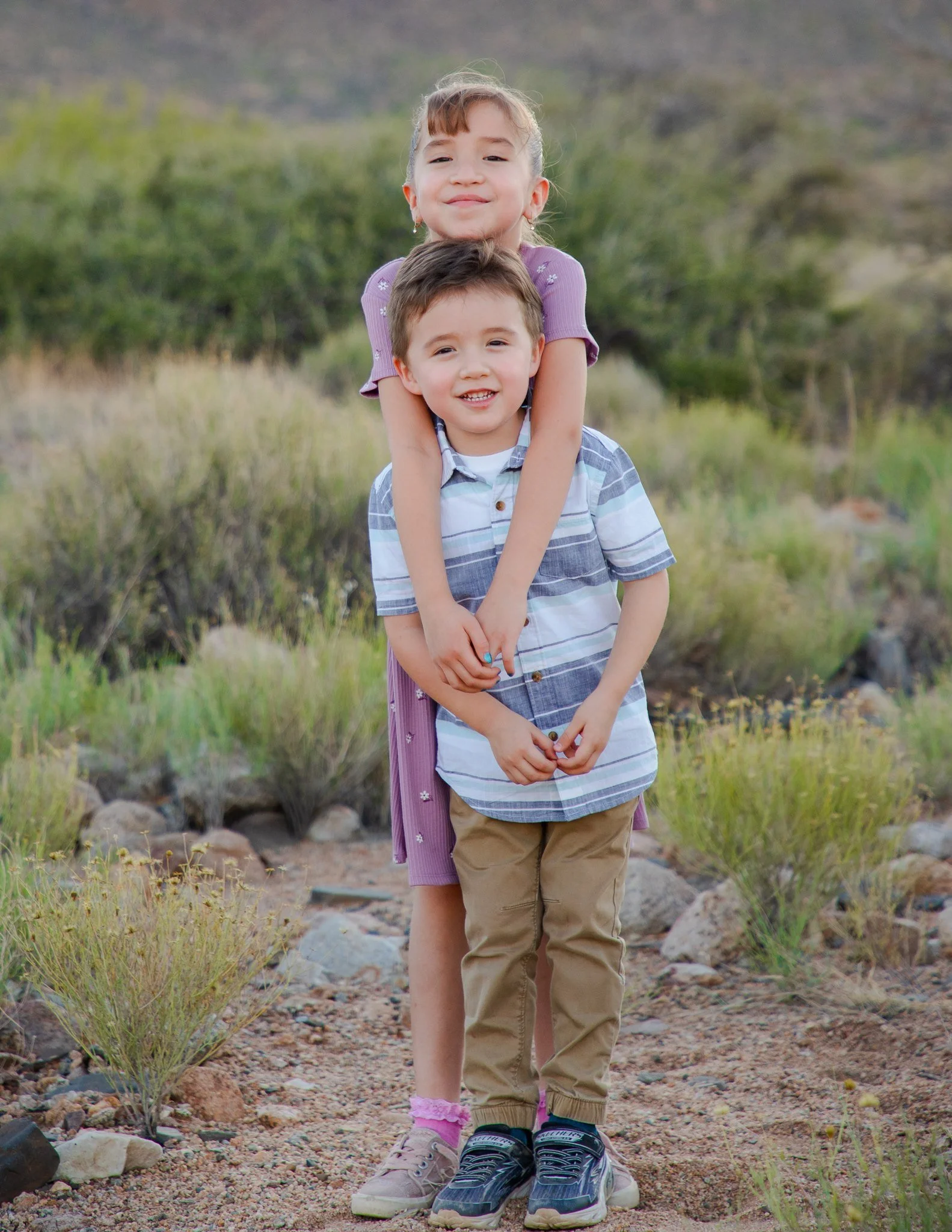 A young girl gives a piggyback ride to a young boy in an outdoor, natural setting with shrubs and rocks, both smiling and standing on a dirt path.