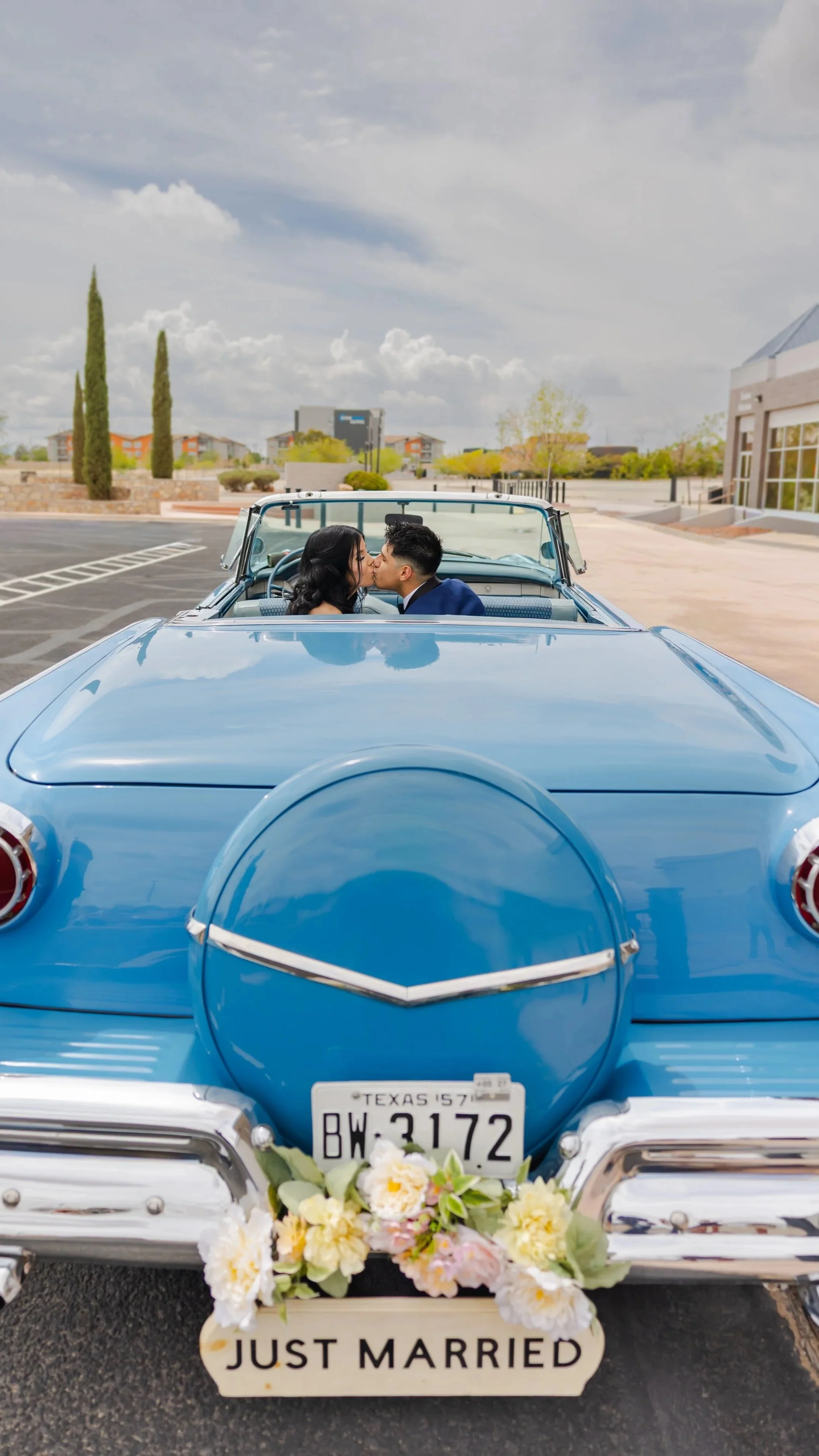 A couple kisses inside a vintage blue car in El Paso, Texas with a Texas license plate, decorated with flowers and a "JUST MARRIED" sign, parked outdoors under a cloudy sky.