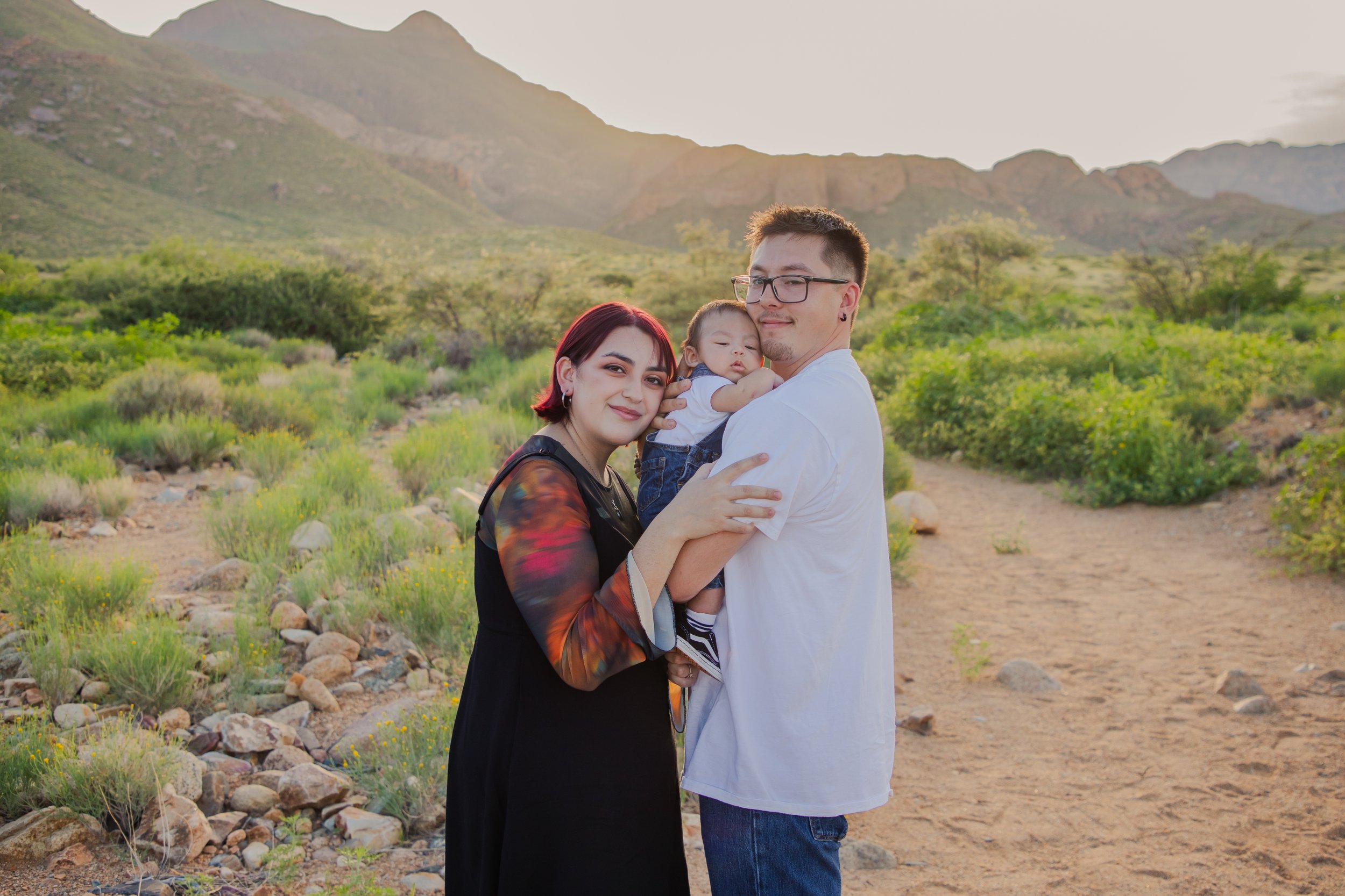 A family of three standing outdoors in a desert landscape with mountains in the background. The woman has short red hair and is wearing a black dress with a multicolored sleeve. The man has glasses, short dark hair, and is wearing a white shirt and blue jeans, holding a baby dressed in denim.