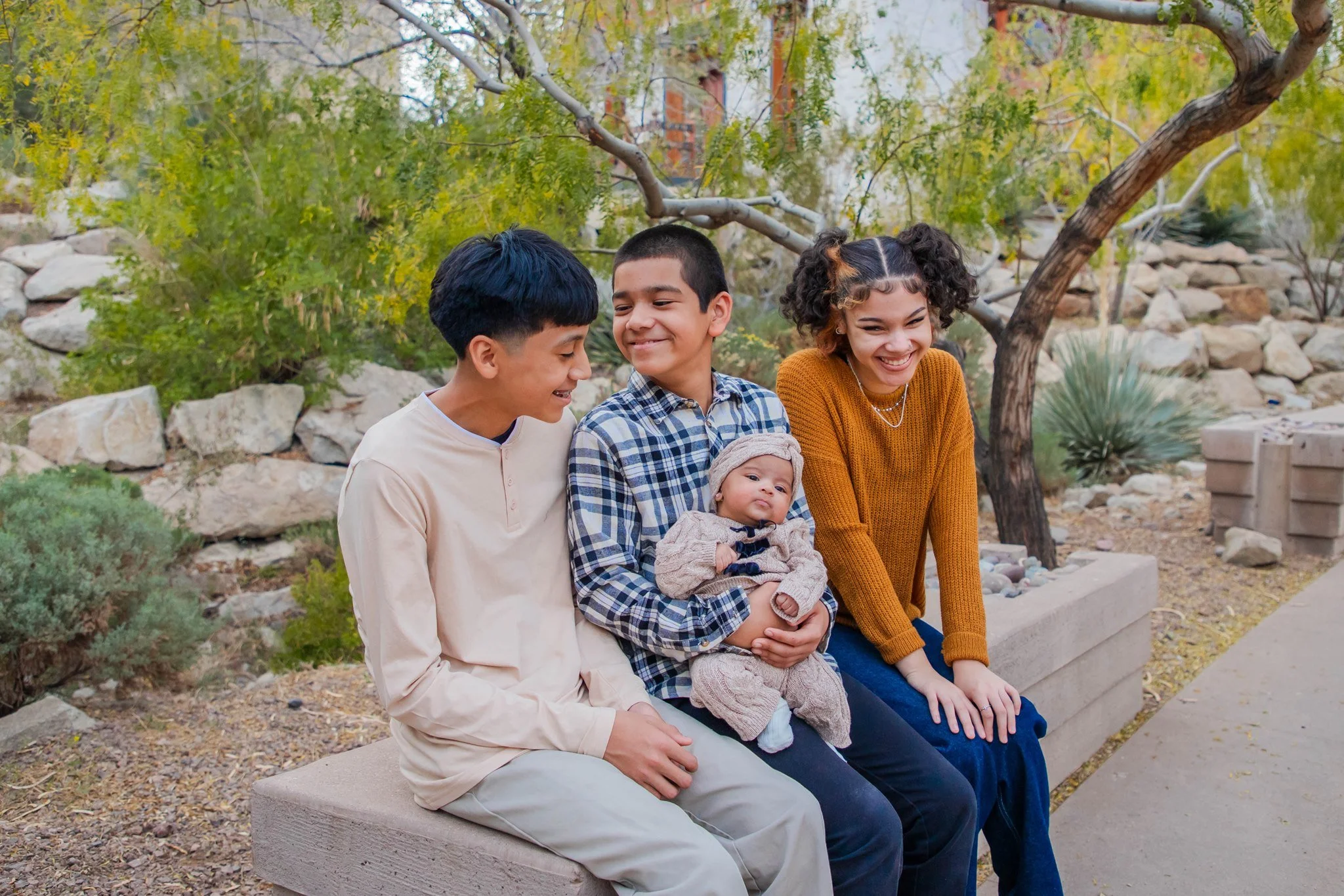 A group of five diverse children sitting on a concrete bench in a park with trees and rocks, all smiling and looking happy.