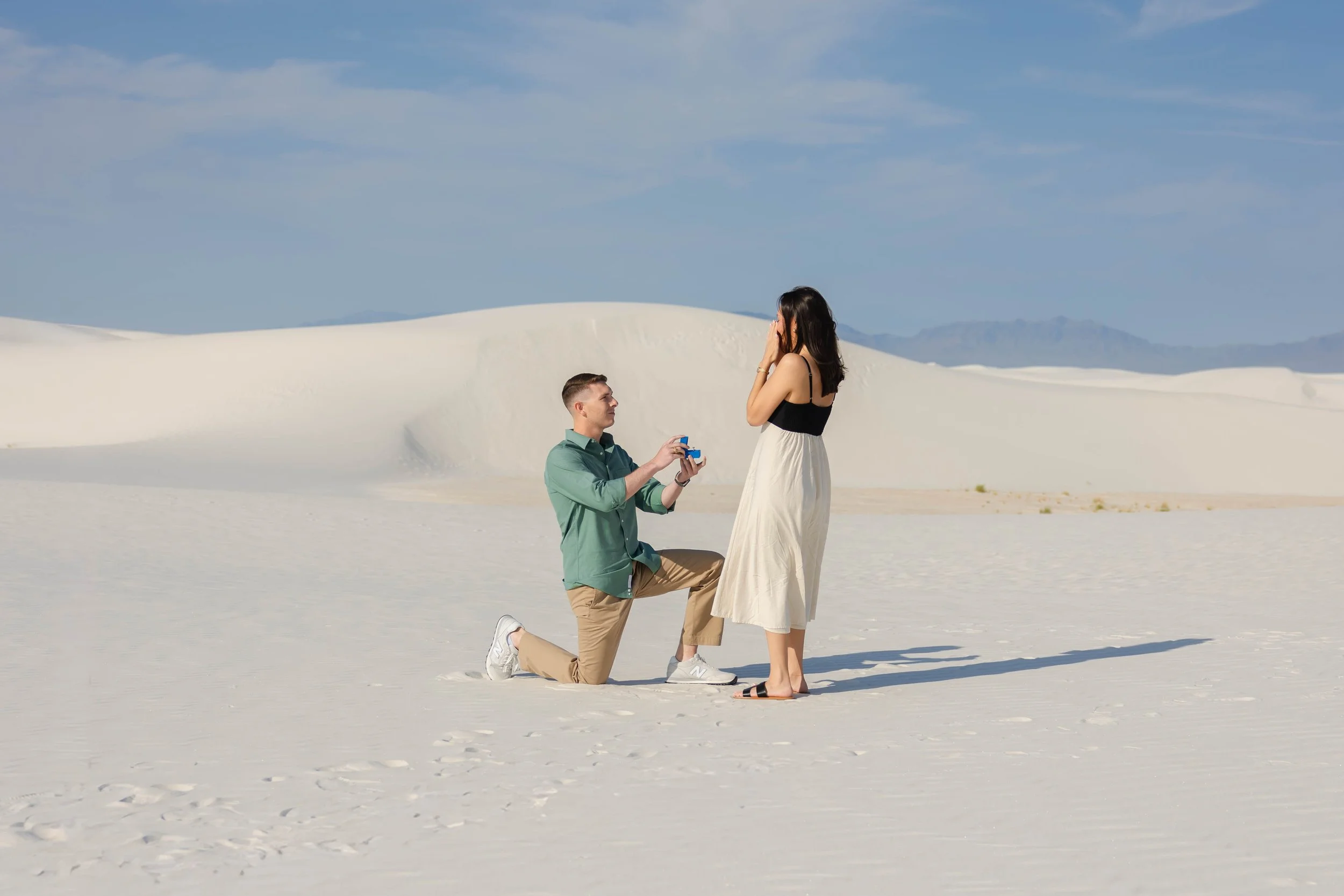 A man kneeling on one knee at White Sands National Park in New Mexico, proposing to a woman who is standing with her hands covering her mouth, both dressed casually, with blue sky and the Organ mountains in the background.