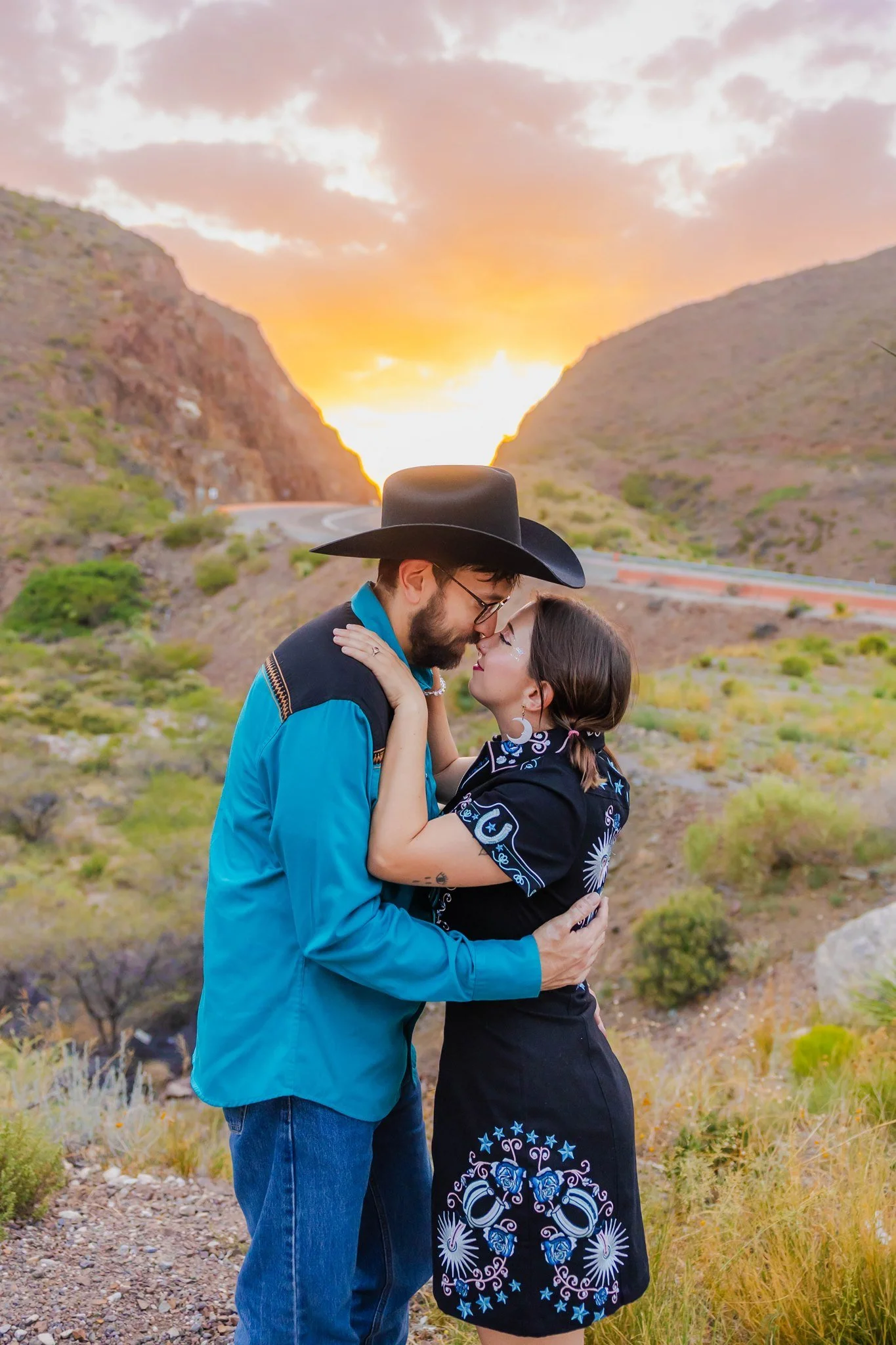 An engaged couple in El Paso, Texas sharing a romantic moment outdoors at sunset by the Franklin Mountains on Transmountain. The man is wearing a cowboy hat and glasses, and the woman is wearing a black dress with blue and white embroidery.